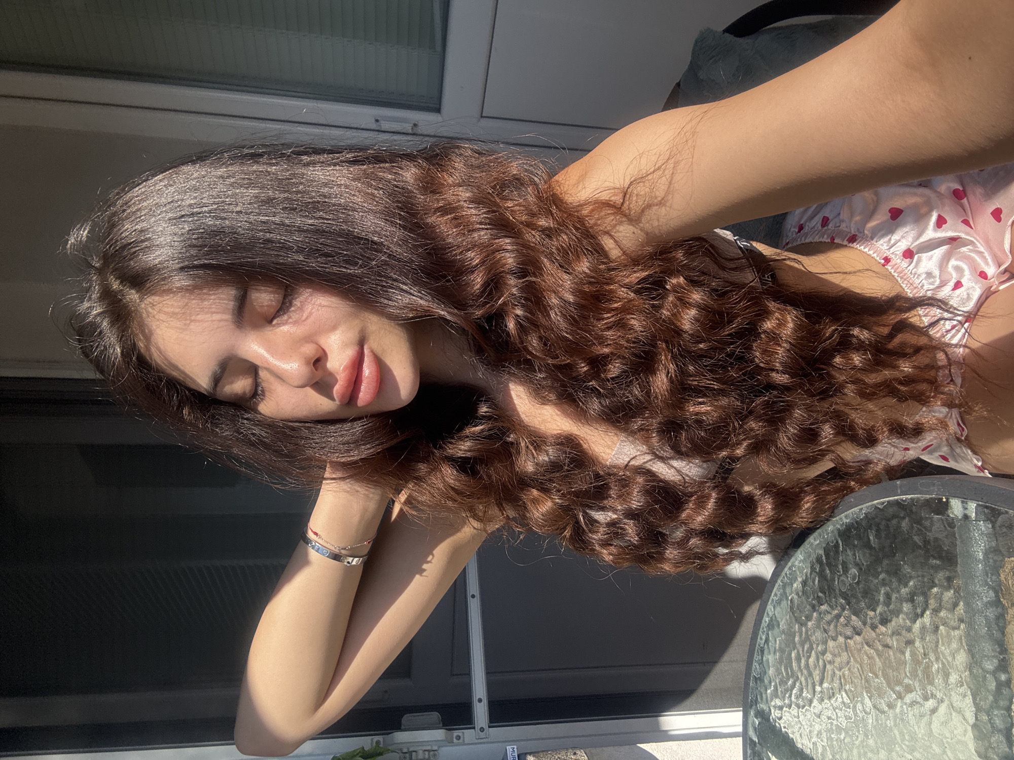A young woman with long, wavy brown hair is posing outdoors, with her eyes closed and her head tilted back slightly. She is wearing a light-colored bralette and pink satin shorts with heart patterns. Her right arm is raised and bent behind her head, and she is wearing bracelets on her left wrist. The background shows a sliding glass door with blinds and a portion of a textured glass table. Sunlight is casting shadows across the scene. The location is identified as Plovdiv, Bulgaria.