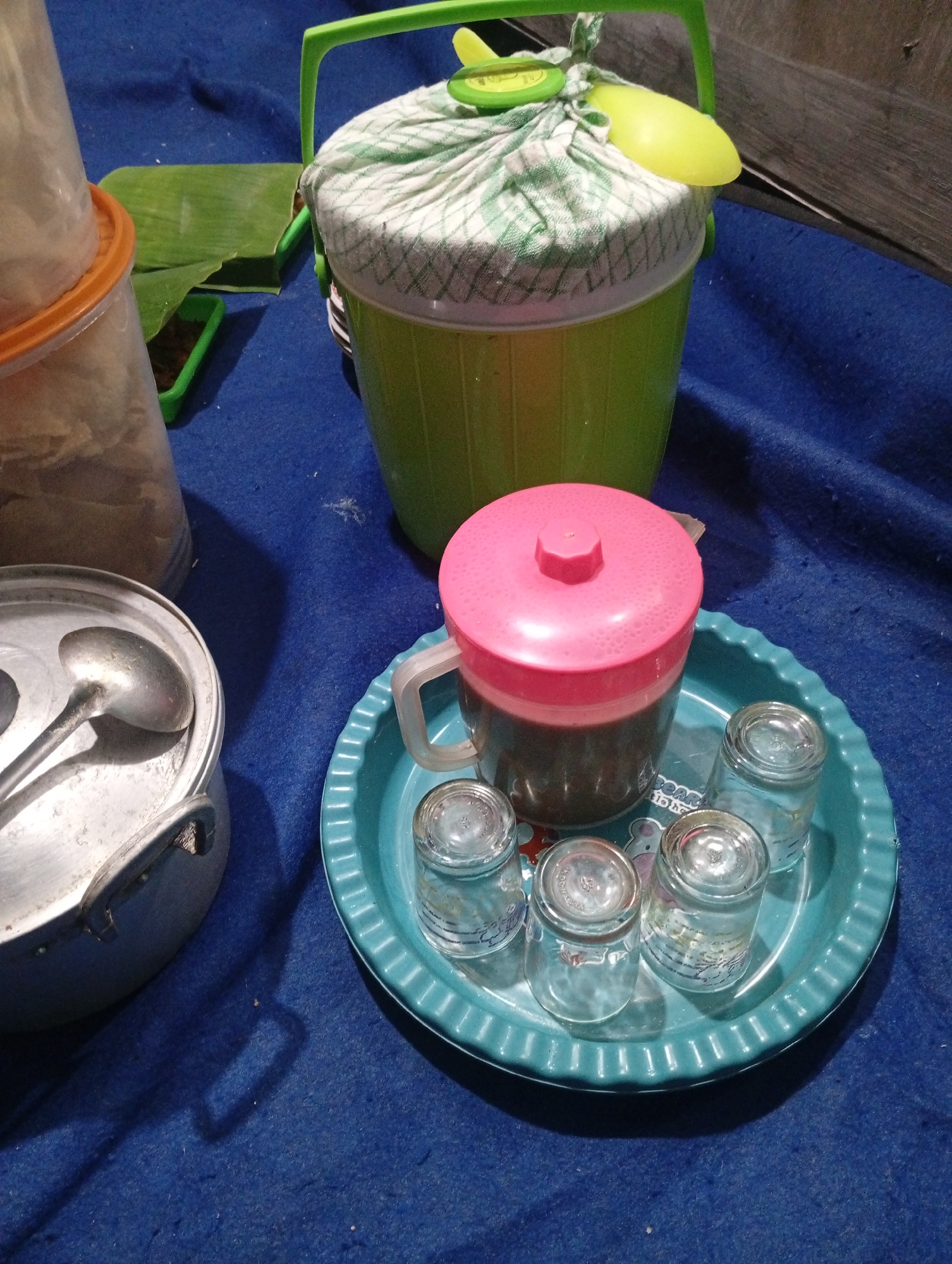 This is a medium shot, eye-level, indoor photograph of a domestic setting, likely a kitchen or dining area, on a blue textured surface. The scene is set in Jatisari, Indonesia.
Dominating the center of the frame is a teal-colored plastic tray with a fluted edge, holding four small, clear glass tumblers and a glass pitcher with a pink lid. The tumblers are empty and appear to have some faint patterned text. The pitcher contains a dark liquid, possibly coffee or tea.
To the left of the tray is a silver-colored metal pot with two handles, and a ladle resting inside it. To the left of the pot, a tall, translucent plastic container with an orange lid is partially visible, containing a light-colored granular substance, likely rice or sugar. Behind this container, a green plastic tray holds a folded banana leaf, and what appears to be a small pile of dark, granular material, possibly spices or coffee grounds.
In the background, a large, lime green bucket with a checkered cloth cover secured by a yellow plastic utensil is positioned on the blue surface. The bucket has a green plastic handle. The overall impression is of a collection of items prepared for serving a beverage or meal. The lighting is dim and appears to be from a single source, casting shadows.