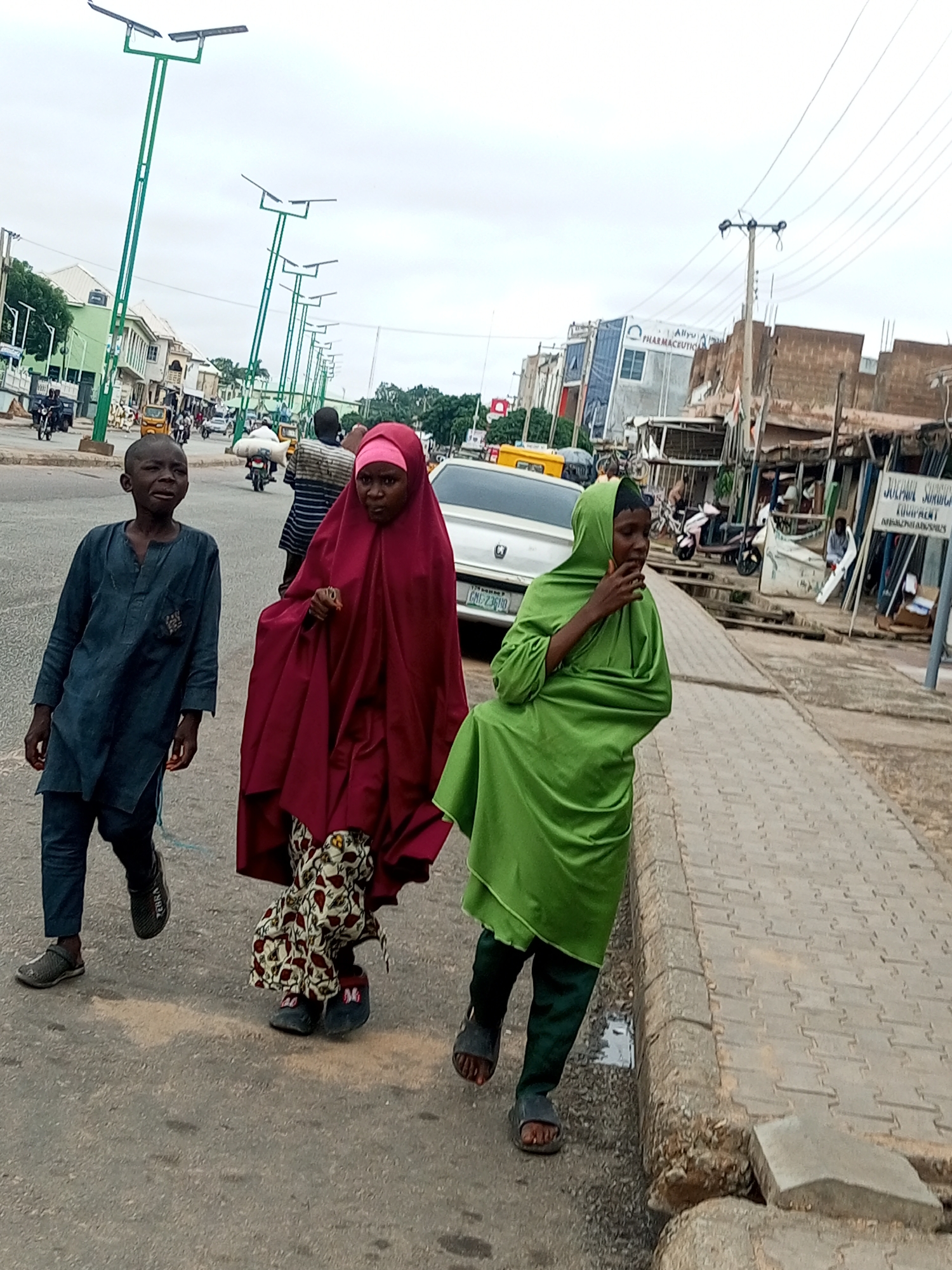 In Arawa, Nigeria, three individuals are walking along a paved sidewalk adjacent to a street. The person on the left is a young boy in a dark blue tunic and pants, with dark shoes. Next to him, a woman in a maroon hijab and matching abaya, with a patterned skirt visible beneath, is walking. To the right, a person in a bright green hijab and pants is walking. They are all moving from left to right in the frame. The street is lined with solar-powered streetlights, buildings, and some market stalls. The sky is overcast. Traffic, including motorcycles and a car, is visible in the background.