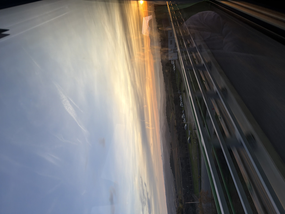 The image captures an outdoor scene at either sunrise or sunset, viewed through a glass panel, possibly from an elevated position. The orientation of the image is rotated 90 degrees clockwise, so for clarity, the description assumes a standard landscape orientation with the sky at the top.
The upper portion of the frame is dominated by a vibrant sky, transitioning from a soft, light blue on the left to intense hues of orange, yellow, and golden light on the right. Faint, wispy clouds or contrails stretch horizontally across the bluer parts of the sky. A bright, intense light source, presumably the sun, is visible on the far right edge, casting a warm glow across the scene.
Below the colorful sky, a distant, hazy landscape extends horizontally across the middle of the image. This landscape appears as dark, undulating hills or low mountains, suggesting a wide expanse of land.
The bottom section of the image reveals human-made infrastructure. Several parallel, horizontal bands, appearing as lanes or structural beams, stretch from the middle-left towards the bottom right. These bands are dark grey and green, suggestive of a multi-lane roadway, railway, or an elevated track or bridge. To the left of these lanes, there's an area of green ground, possibly grass, where several vehicles (likely cars) are parked in a row. Further back from the parked cars, a few small, boxy white buildings are visible.
Numerous reflections are present on the glass surface through which the image is captured. In the upper-right area of the sky, a reflection of a white, rectangular object with some dark text or a logo is visible, resembling a flag or banner. In the lower-right quadrant, superimposed over the infrastructure and horizon, a distinct reflection of a person's upper torso and arm is visible. The person appears to be wearing a light-colored, soft, or fluffy garment, possibly a robe or blanket. The reflections of the buildings and parked cars also appear slightly ethereal or layered with the actual view, indicating the presence of glass.
The warm, golden light and the sun low on the horizon strongly suggest the time of day is either sunrise or sunset. No specific text is clearly legible, though text is hinted at on the reflected white banner.