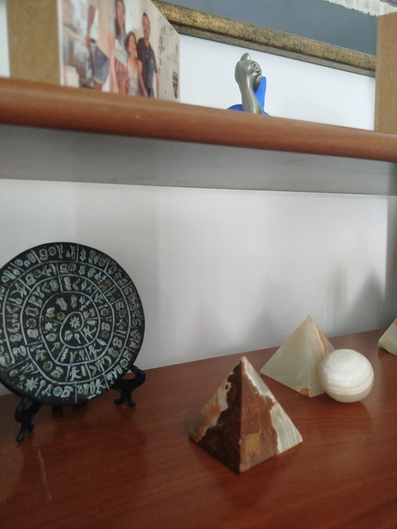 A close-up, eye-level shot shows a wooden shelf with various decorative objects. On the left, a circular, dark-colored object with intricate carvings and symbols resembling ancient script rests on a black stand. To the right of this object are two stone pyramids and a spherical stone. The larger pyramid in the foreground has a variegated brown and white pattern, while the pyramid behind it is translucent and pale green. The sphere is white and marbled. In the background, a framed photograph of a family is visible on an upper shelf, along with a small metallic figurine of a cat. The overall setting appears to be an indoor display shelf. The location in the city of Ashkelon, Israel, cannot be determined from the image alone.