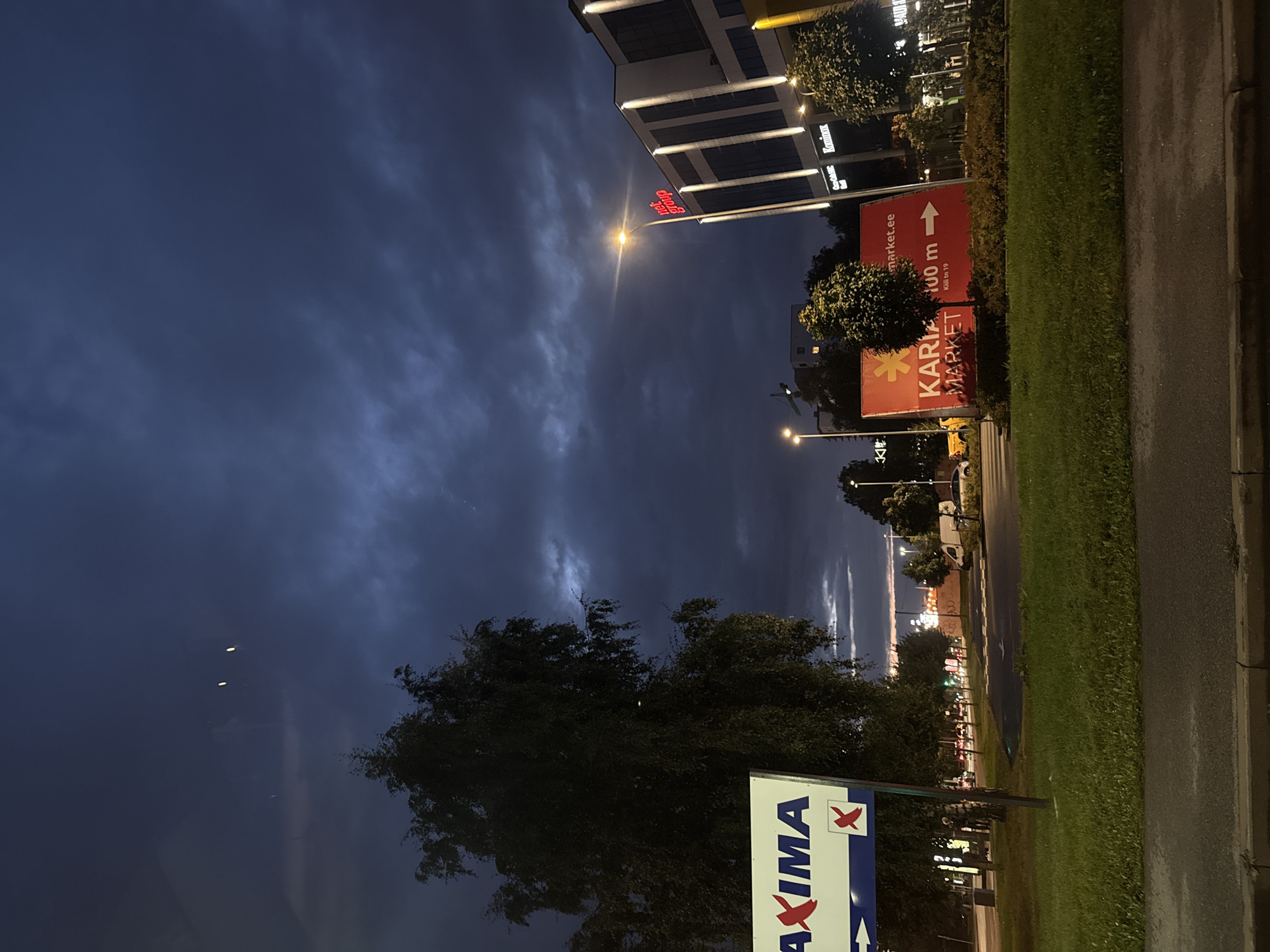 The image depicts a street scene in Tallinn, Estonia, during twilight or early night. The sky is dark with dramatic cloud formations, illuminated by distant lights. On the left, a large sign for "MAXIMA" is visible, a popular supermarket chain in the Baltic region. Next to it is a tall, leafy tree.
Further to the right, a modern building complex is illuminated, with the prominent red signage "net group" on its facade. Below this, smaller signs like "On24.ee" and "Kari market" are partially visible, indicating businesses within the complex. A street lamp casts a bright light on the building and the surrounding area.
In the foreground, a red sign with white text announces "KARIA MARKET", with an arrow pointing to the right and indicating "100 m". The scene includes a well-maintained grassy area and a paved road, with a few cars and a white van visible in the background. The overall impression is of a commercial area at dusk, with artificial lights beginning to dominate the natural light.