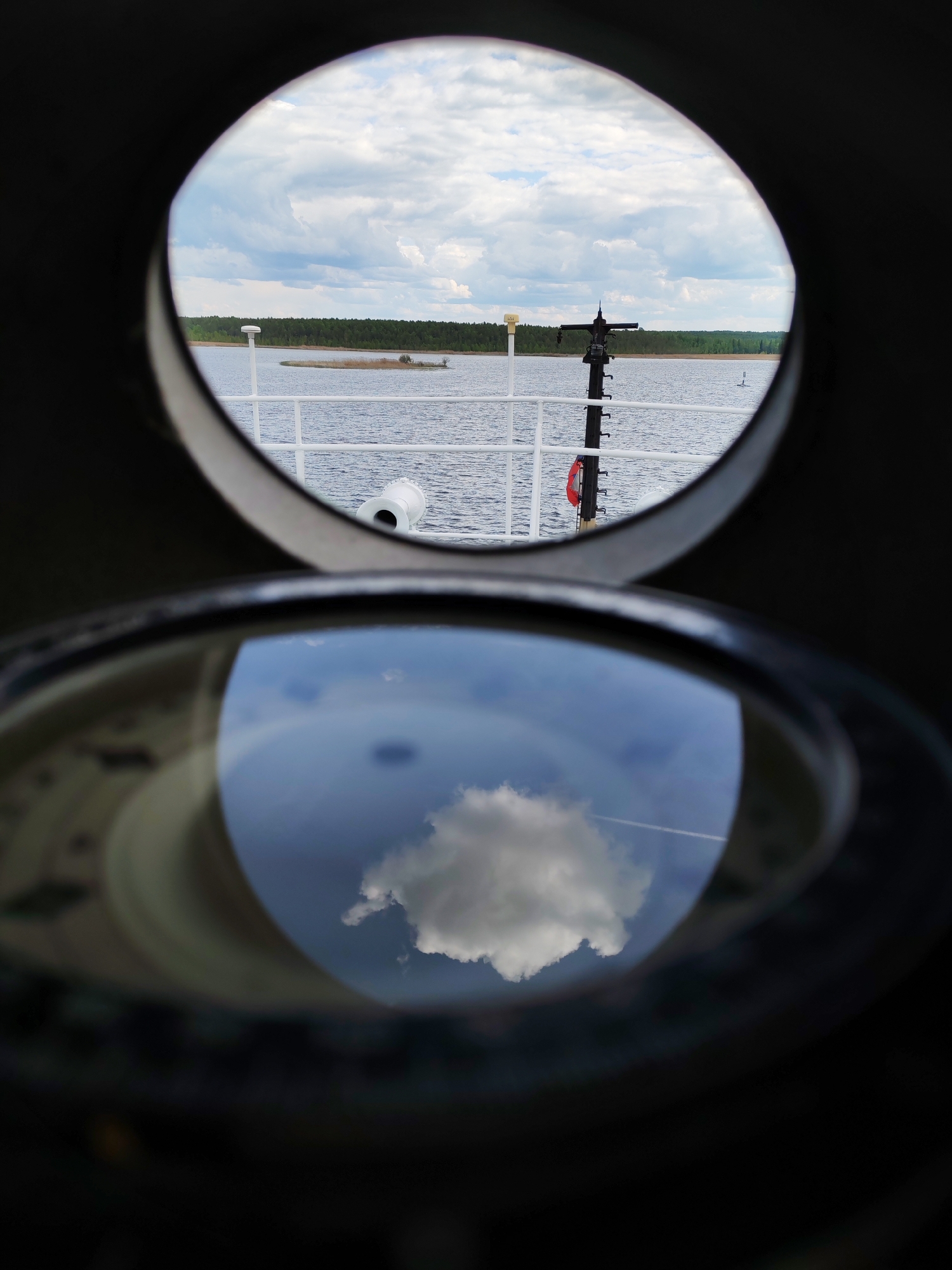 A porthole-style window and a compass-like dial are in the foreground, with a view of a body of water, land, and cloudy sky visible through the window. The reflection in the dial shows a cloud in the sky and a contrail. A white railing, a white object, a black pole, and a red life preserver are visible through the window. The location is likely a ship in Novodevich'e, Russia.