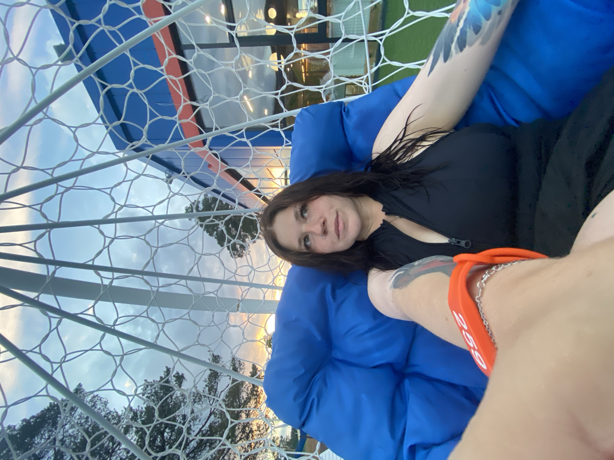 A young woman with dark hair and piercings sits in a blue beanbag chair within a white geodesic dome structure at an outdoor venue. She is wearing a black swimsuit and an orange wristband with the number "259" visible. The background shows a blue building with red accents, trees, and a cloudy sky with sunset hues. The image appears to be taken in Votkinsk, Russia.