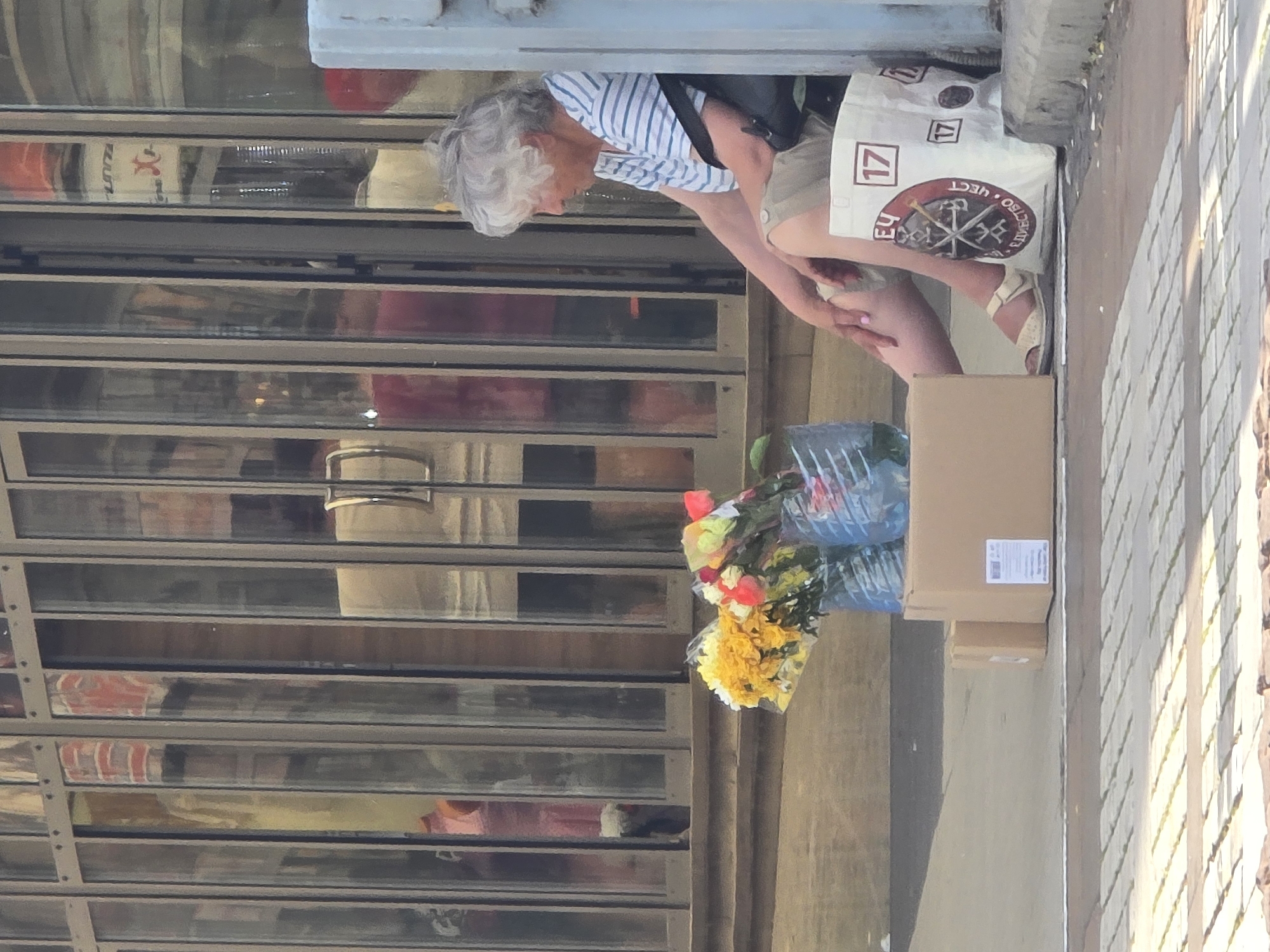 The image shows a woman with gray hair, wearing a blue and white striped shirt and khaki shorts, sitting next to a building with glass doors. She is next to a cardboard box, flowers wrapped in blue plastic, and a white bag with a logo and the number 17. The location appears to be a city street, but the specific city is unknown. The woman is looking down at her hands. The image is oriented sideways.