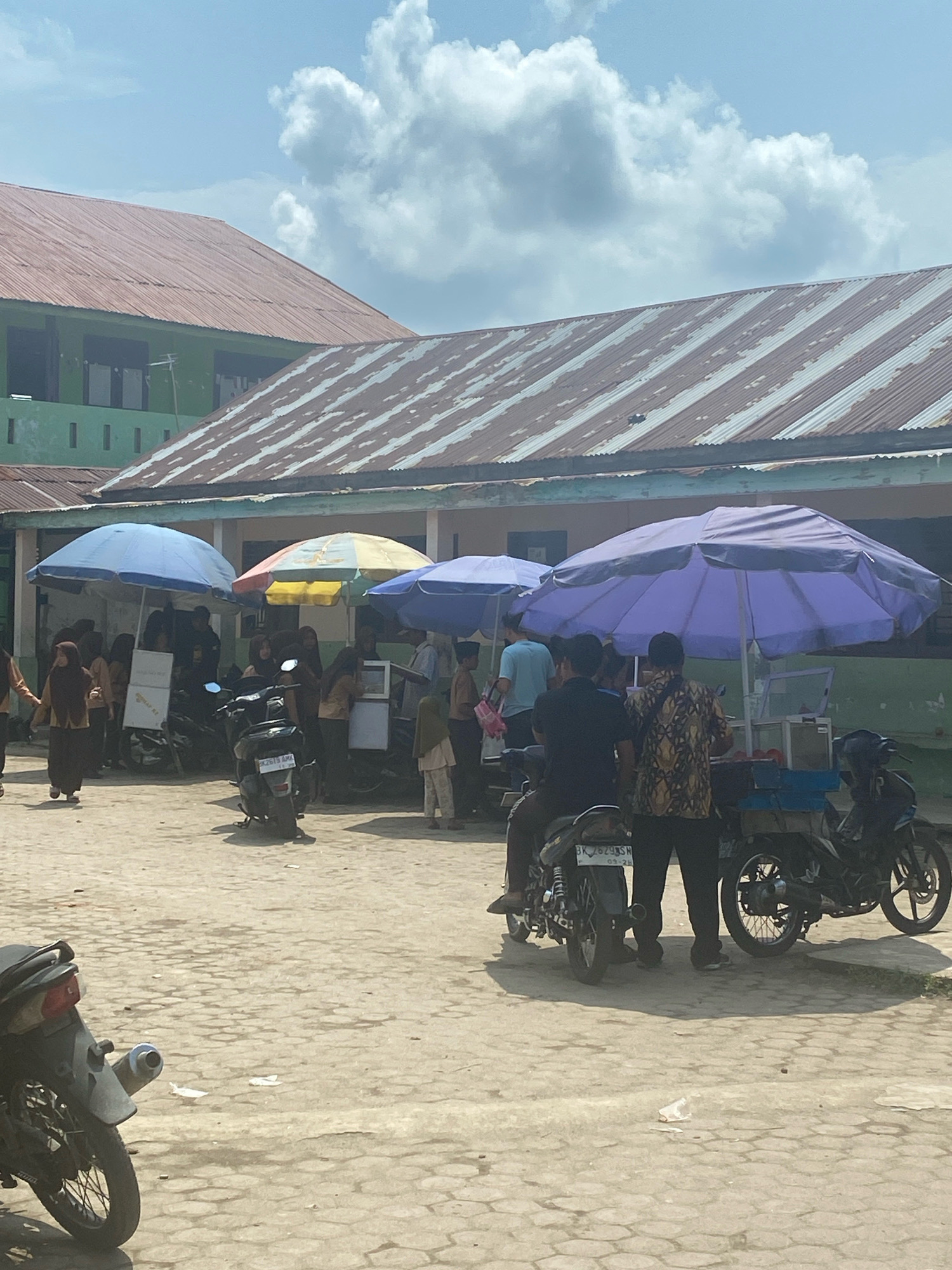 The image depicts an outdoor scene with several food stalls set up under large umbrellas in front of a building with a corrugated metal roof. The stalls are mobile, mounted on the backs of motorcycles. People, likely students, are gathered around the stalls, some wearing brown uniforms and headscarves. The ground is paved with interlocking concrete blocks. In the foreground, a motorcycle is parked. In the background, the building appears to be a school or institutional building with a green facade. The sky is partly cloudy. The scene appears to be a bustling marketplace or gathering area, likely near a school in Karang Gading, Indonesia.