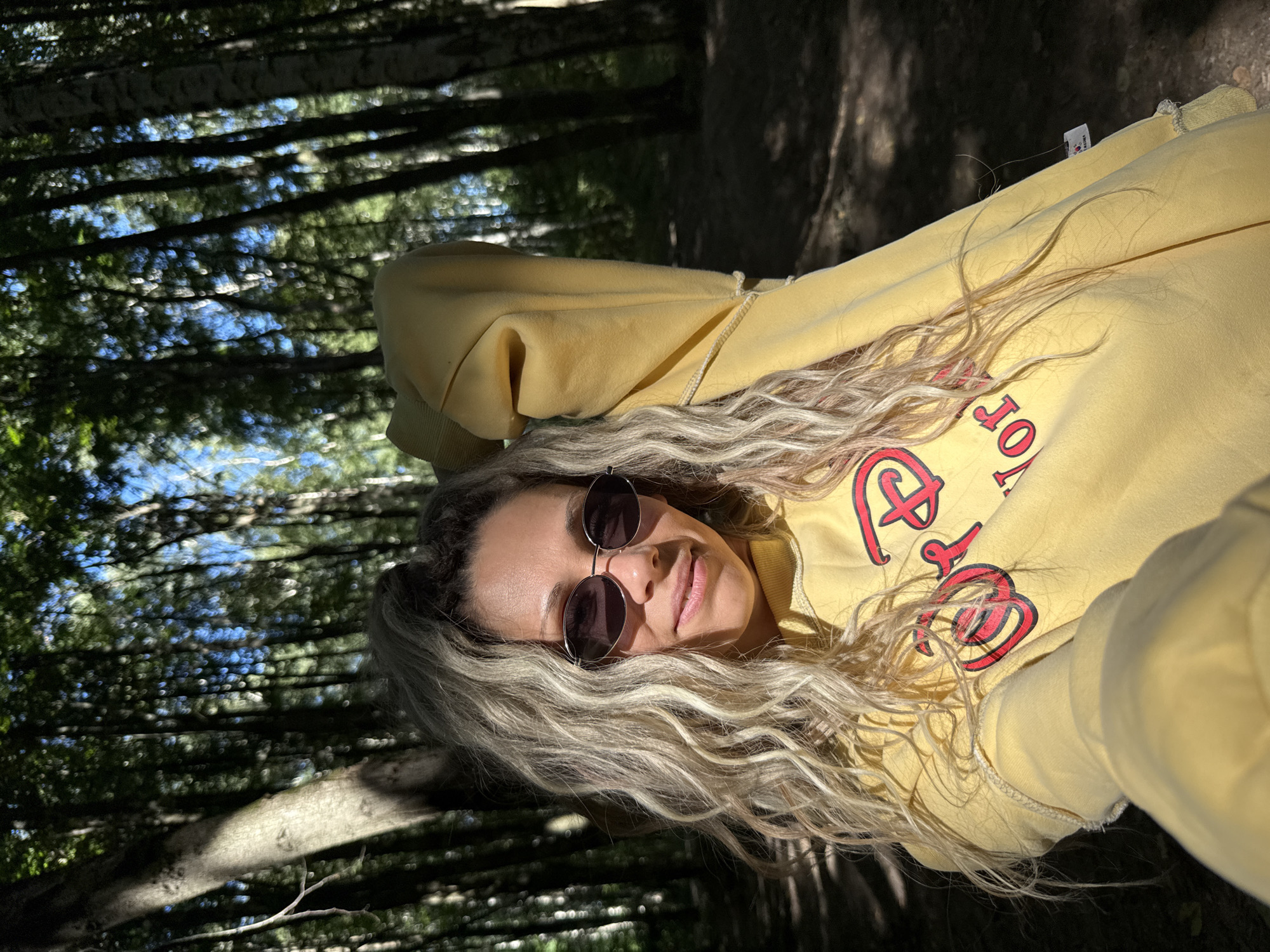 A woman with blonde, curly hair and sunglasses is taking a selfie in a forest. She is wearing a yellow sweatshirt with a red and black graphic of "Disney World" on it. The background is a dense forest with tall trees and dappled sunlight. The image appears to be taken in the city of Rusanovo, Russia, given the context of the prompt.