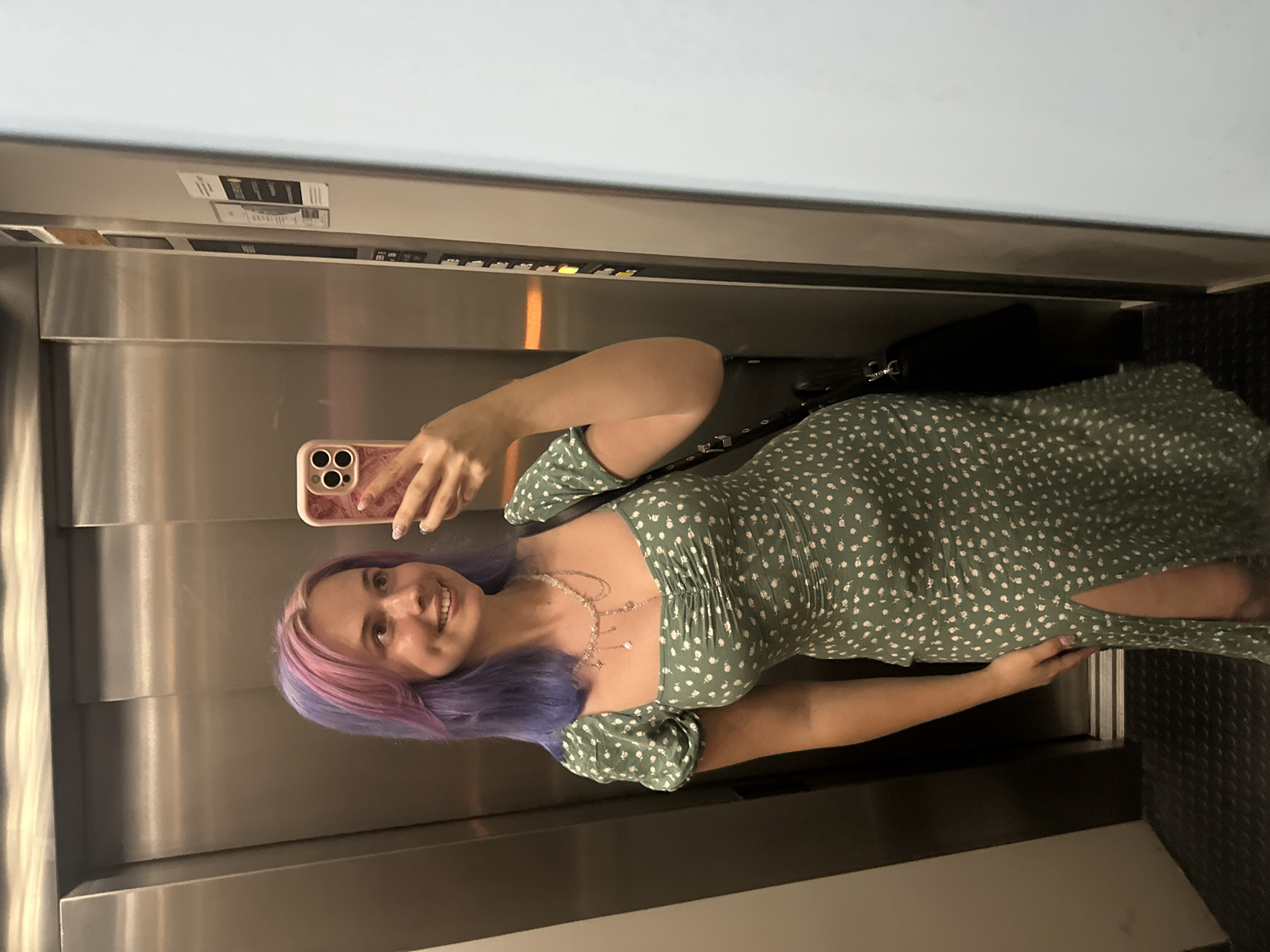 A woman is taking a selfie in an elevator. The woman has dyed hair, with purple and pink colors. She is wearing a green floral dress and a necklace. She is holding a pink cell phone. Behind her is the stainless steel interior of the elevator and a control panel. The exact location in Gijón/Xixón, Spain is impossible to determine based solely on this image.