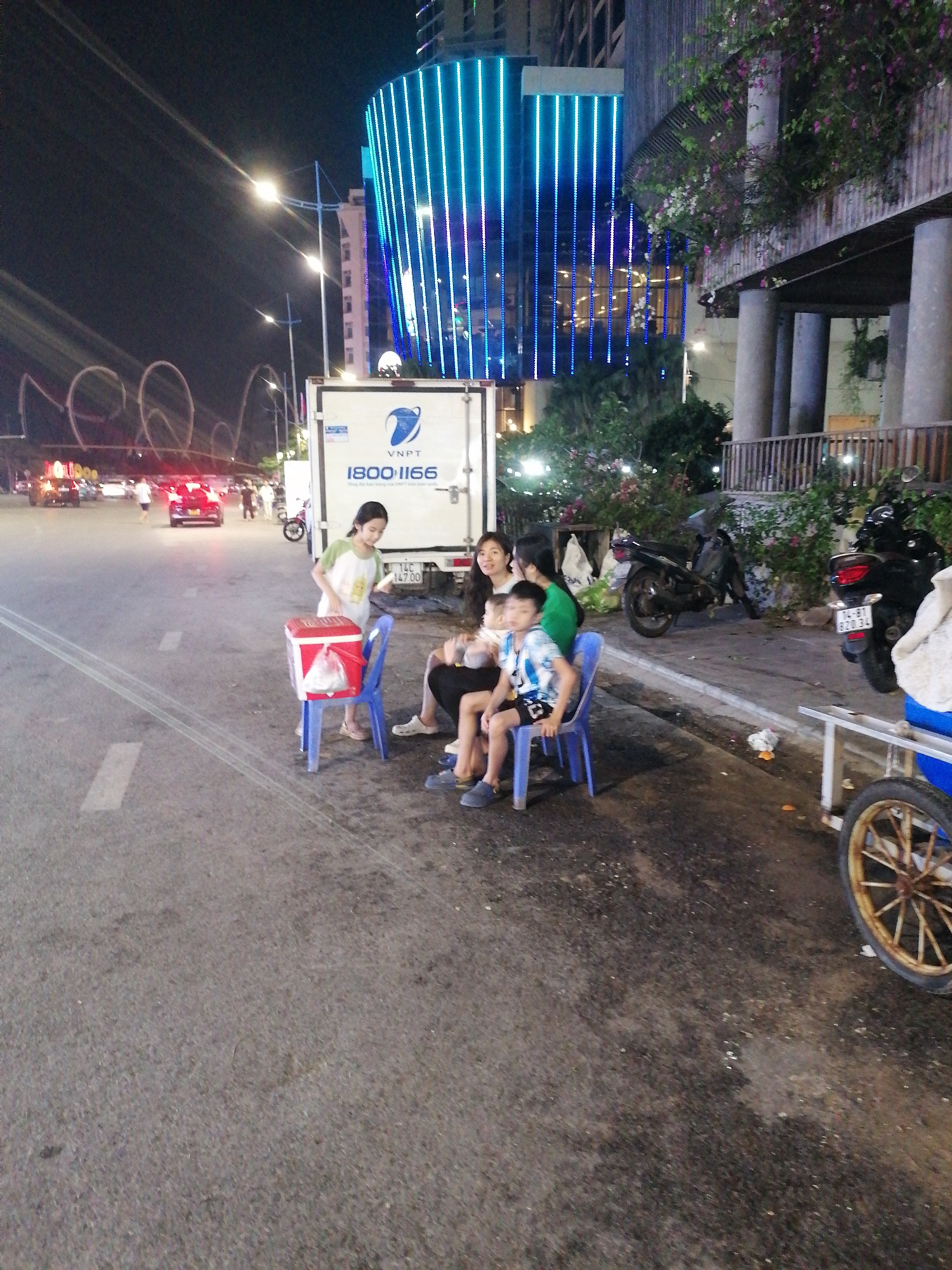 In Bai Chay Ward, Vietnam, a family is sitting on blue plastic chairs next to a road at night. To the left, a red and white cooler sits atop another blue plastic chair. A girl stands nearby, possibly tending to the cooler. Behind them is a white truck with the logo of VNPT and the phone number 1800-1166. Further back, a street is visible with cars and motorbikes passing by. In the background is a tall building with vertical blue lights. To the right, there is a sidewalk with parked motorbikes and a cart with a wheel. The scene is lit by streetlights and the lights from the building.
