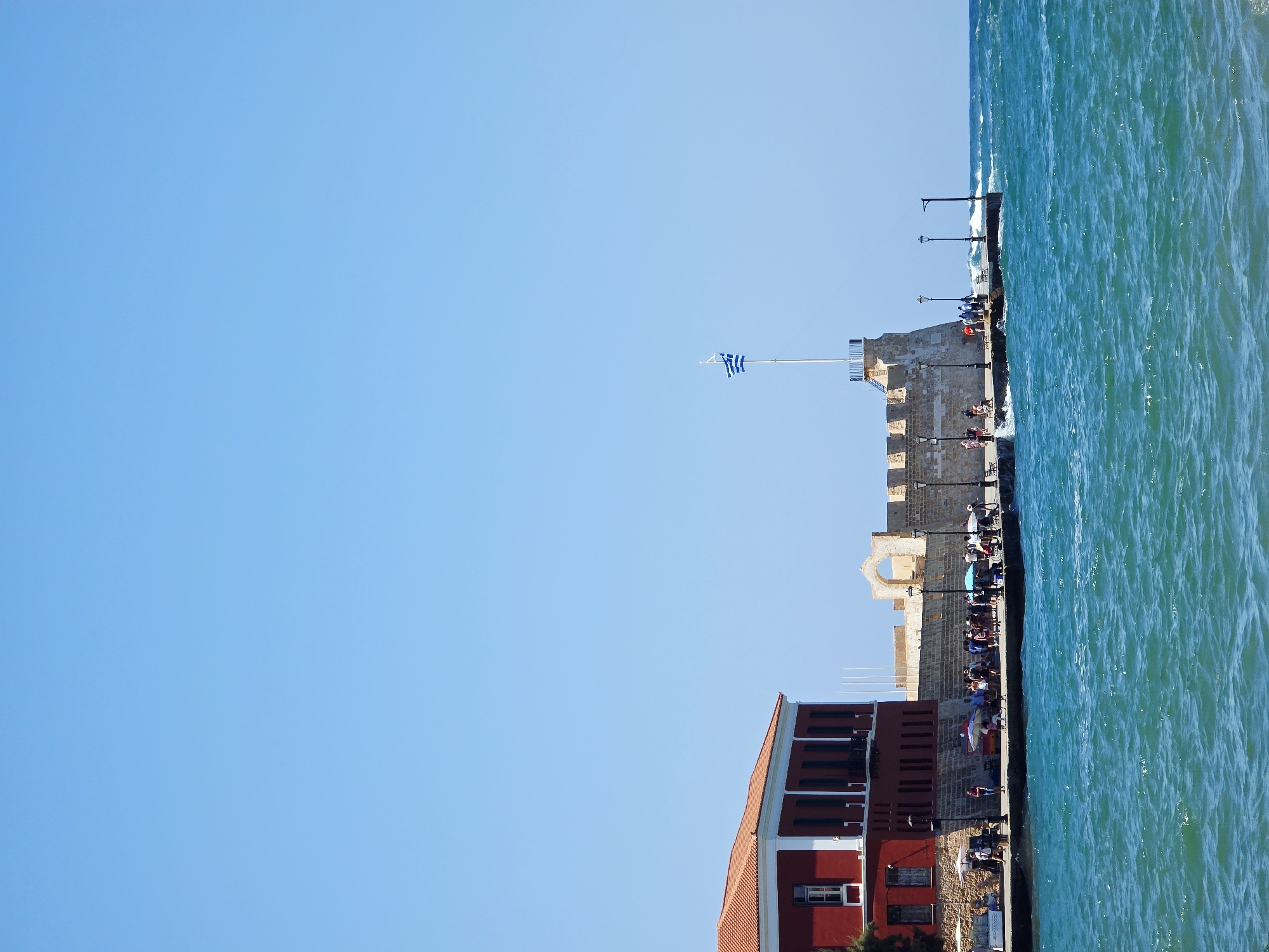The image depicts the Venetian harbor in Chania, Greece, on a clear, sunny day. In the foreground, the turquoise water of the sea is choppy with visible waves. Along the waterfront, a stone promenade is lined with people walking, sitting, and observing the surroundings. To the left, a prominent red building with a white trim and orange-tiled roof stands, featuring several windows and a balcony. Adjacent to it, a fortified stone structure, likely a section of the old Venetian walls, extends along the harbor. A flagpole with a Greek flag is prominently displayed on top of this fortification. Lampposts are spaced along the promenade. The sky is a vast, cloudless expanse of light blue. The overall scene captures a bustling waterfront area, suggesting a tourist destination.
