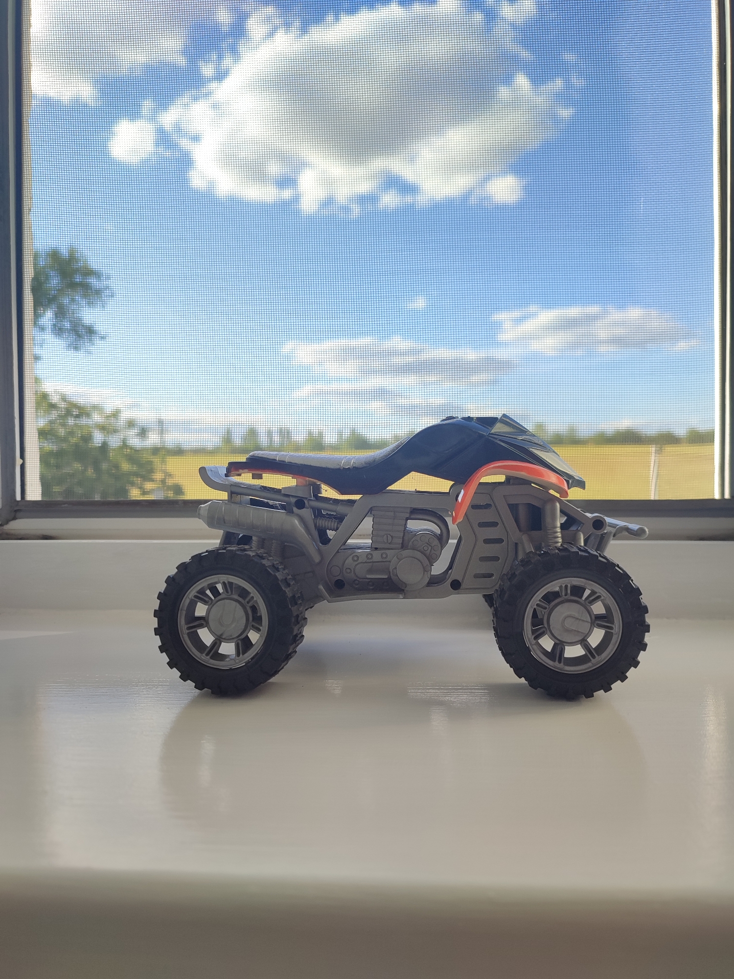 A toy ATV is positioned on a white windowsill, in front of a window. Through the window, a blue sky with scattered white clouds is visible, as well as trees and a yellow field in the distance. The ATV is gray and orange with black tires.