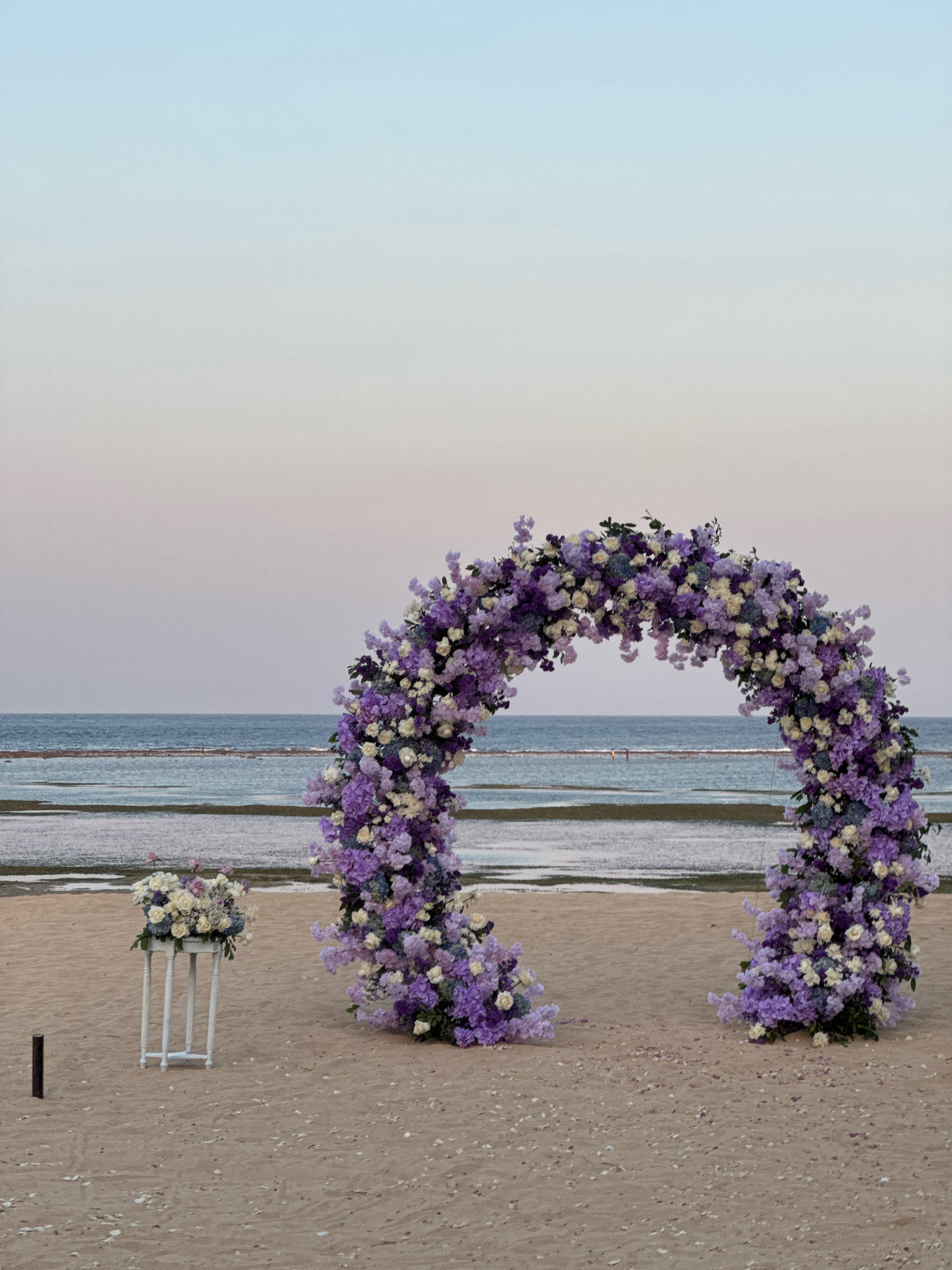 A floral archway, adorned with purple and white flowers, stands on a sandy beach in Kuta Selatan, Indonesia. The arch is situated near the water's edge, with the ocean visible in the background. To the left of the arch, a white pedestal holds a bouquet of white and purple flowers. The sky above is a soft gradient of blue and pink, suggesting either sunrise or sunset. The sand is dotted with scattered shells and debris. No people are clearly visible in the frame.