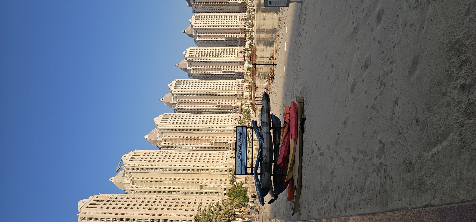The image captures a waterfront scene with a row of identical, light-colored buildings lining the background. The buildings appear to be apartments or hotels, each featuring a distinctive domed roofline and numerous windows. In the foreground, the sandy beach is occupied by a rack holding several kayaks of various colors, including blue, orange, red, and yellow. A sign with blue text on a white background is attached to the kayak rack, reading "Kayak Rack". Palm trees are visible between the buildings and the beach. The sky is a clear, bright blue, suggesting a sunny day. The lighting indicates it is likely late afternoon or early morning, with the sun casting shadows. The overall setting is a modern urban beach environment, possibly a resort area in Doha, Qatar, given the architectural style. There are no people visible in the image.