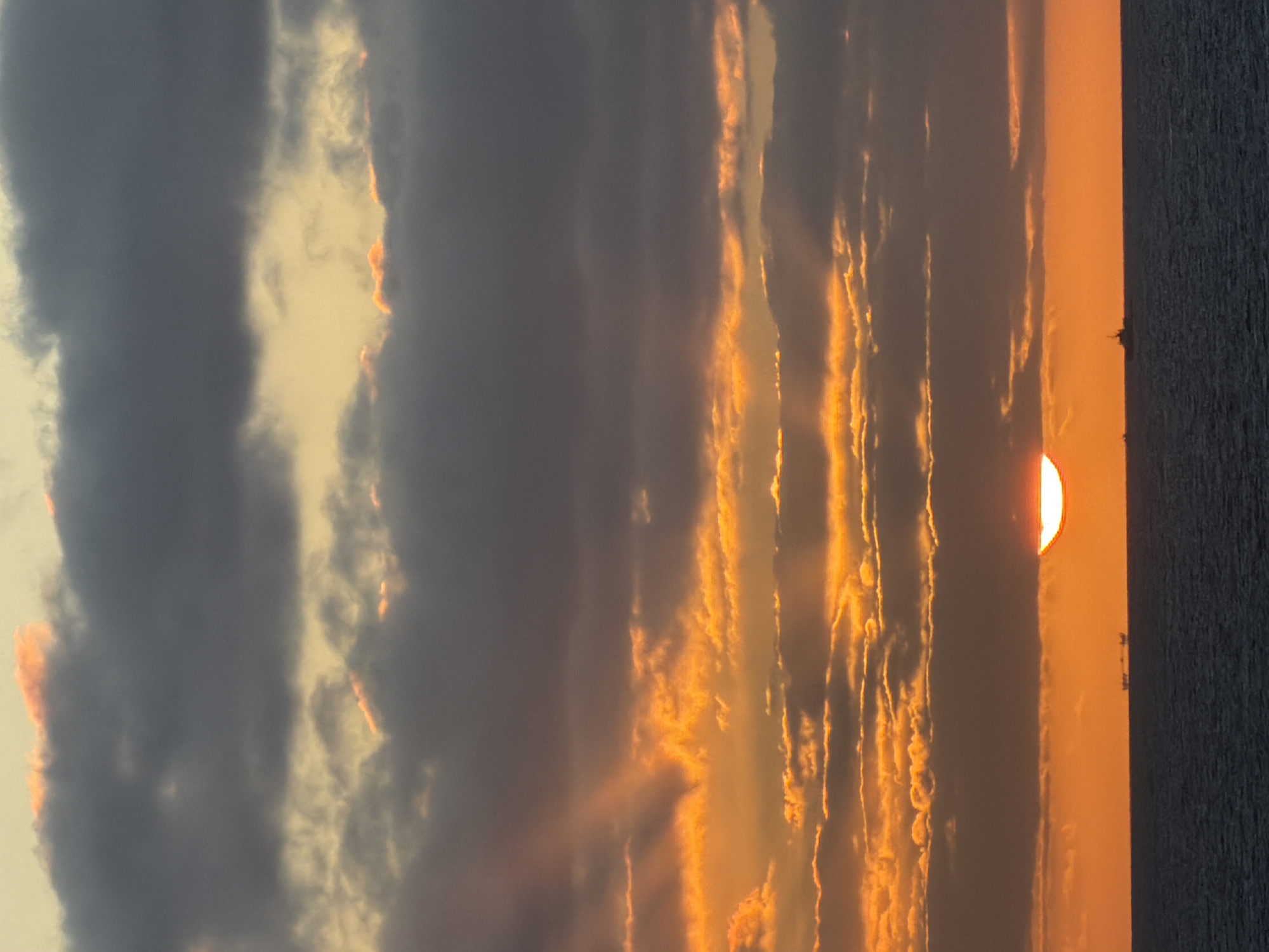 A vertical oriented landscape photograph depicts an atmospheric sunset over water, possibly an ocean, in Unknown, Unknown. The sun, appearing as a semi-circular, bright orange orb, is partially obscured by dark clouds at the water's edge on the right side of the image. The sky is filled with alternating bands of dark grey and orange clouds that run vertically from the top to the bottom of the image. Light beams stream down through some of the breaks in the clouds, creating an ethereal effect. The water on the right is a dark, textured expanse. Small, indistinguishable objects that may be boats or other watercraft, are silhouetted against the bright horizon.