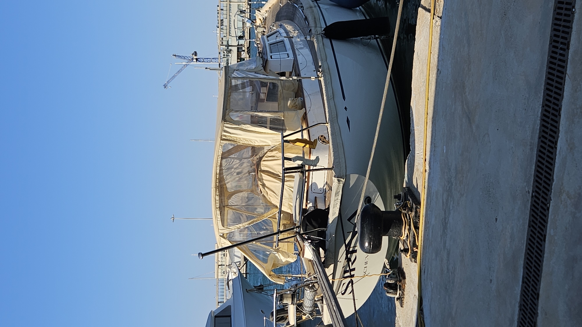 The image shows a side view of a moored sailboat, possibly in Dénia, Spain, due to the context provided. The boat is light-colored with a tan canopy covering its deck and cockpit area. The canopy appears to be made of a translucent material, allowing some light to filter through. There are black railings along the sides of the deck. A large black cleat is visible near the stern of the boat, with yellow and white ropes attached. Another boat, lighter in color, is docked alongside.
In the background, the sky is a clear, bright blue, suggesting a sunny day. A distant construction crane is visible on the left side of the frame, hinting at ongoing development or construction in the port area. The edge of a concrete dock is on the right, with a dark grate running along its length.
No people are visible in the image. The scene appears calm and still, typical of a marina on a fair day. The time of day seems to be daytime, judging by the bright sunlight and clear blue sky. The overall impression is one of a peaceful harbor scene. There is some text visible on the stern of the sailboat, but it is partially obscured and difficult to read definitively.
