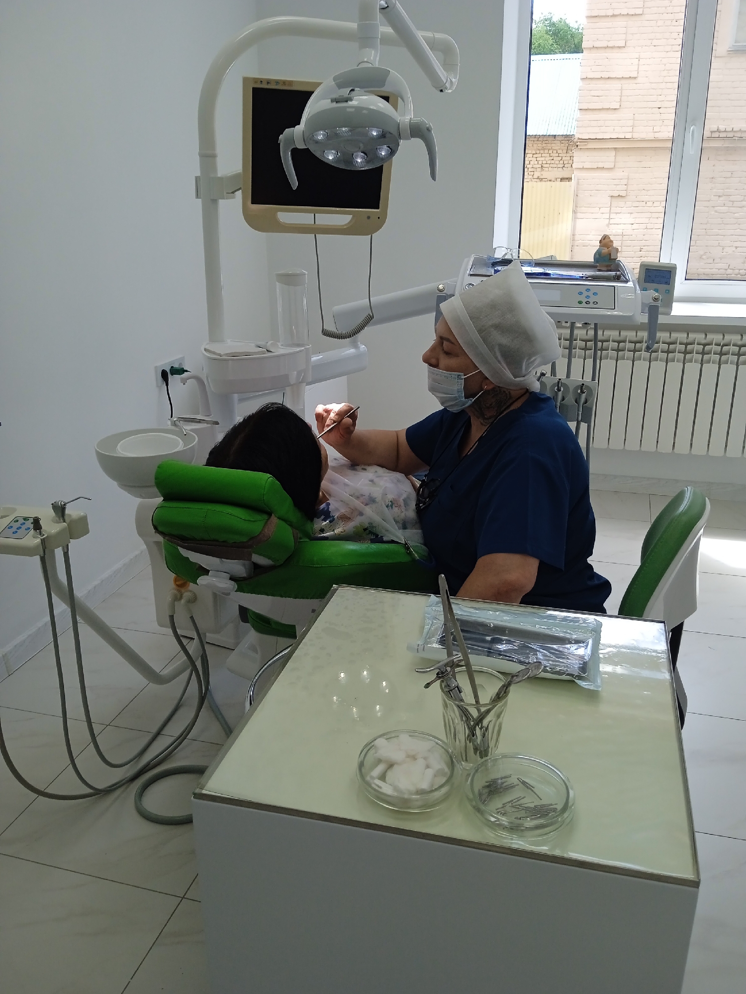 A dentist, wearing a blue scrub top and a white surgical cap and mask, is examining a patient in a dental clinic. The dentist is leaning over the patient, who is reclined in a green dental chair. The dentist is holding a dental instrument in her right hand, which is near the patient's mouth. The patient's head is on a green headrest. To the left of the patient is a dental unit with a sink and various instruments. In the foreground, there is a light-colored table with dental supplies, including cotton balls, pins, and instruments in a holder. The clinic appears to be modern, with white walls and tiled floors. A window in the background looks out onto a brick building and trees. The location is identified as a dental clinic in the city of Oral, Kazakhstan.