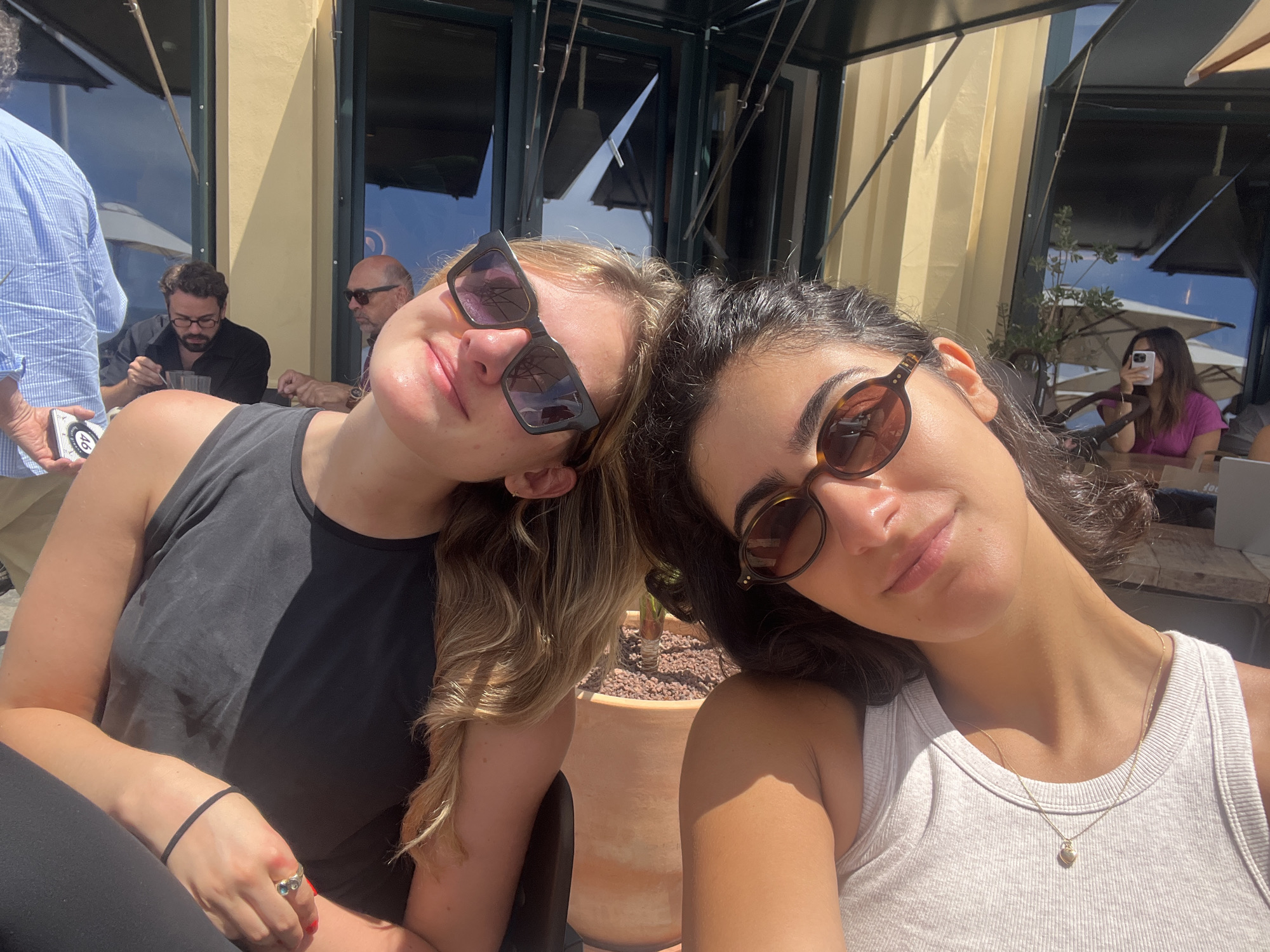 Two young women are posing for a selfie outdoors, their heads tilted together. They are seated at tables in front of a restaurant with large glass windows and awnings. The woman on the left is wearing a black tank top and sunglasses. The woman on the right is wearing a white ribbed tank top and sunglasses. In the background, several other people are visible, some seated at tables, and one person is taking a photo with a phone. The environment appears to be a sunny day. The presence of specific restaurant signage or architectural features that definitively link the location to Porto, Portugal is not present in the image.