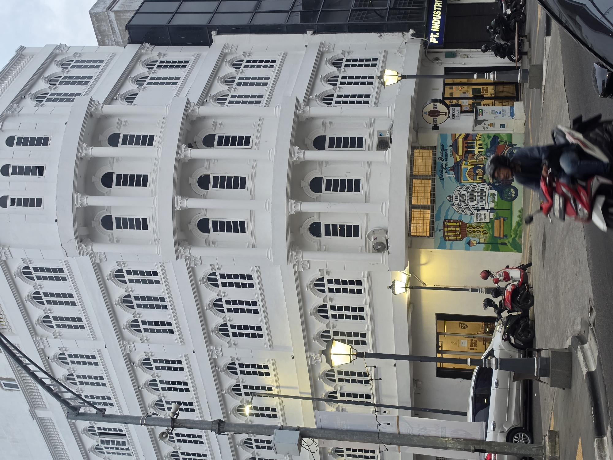 This is a vertical-format, eye-level shot of a white, multi-story building with numerous windows. The building's facade features architectural details like columns and decorative elements above some of the windows. The windows themselves are large and rectangular, with some arched tops. A street runs in front of the building, with a sidewalk.
Several objects are visible on the street and sidewalk. A white car is parked on the right side of the frame, and two motorcycles are parked behind it. A person is riding a red and white motorcycle in the foreground, partially obscuring the building. Streetlights are visible, one of which is illuminated, suggesting it might be late afternoon or evening. A mural is painted on the lower part of the building's wall, depicting several buildings.
Text visible in the image includes "HERITAGE MEDAN," "Heritage Boutique HOTEL MEDAN," "BY TONGA," "BREW BEAN COFFEE SHOP," and "PT. INDUSTRI." The presence of "HOTEL MEDAN" suggests the building may be a hotel. The overall scene appears to be in a city, likely during daylight hours, though the illuminated streetlight suggests it could be nearing dusk or early morning. The weather appears to be overcast.