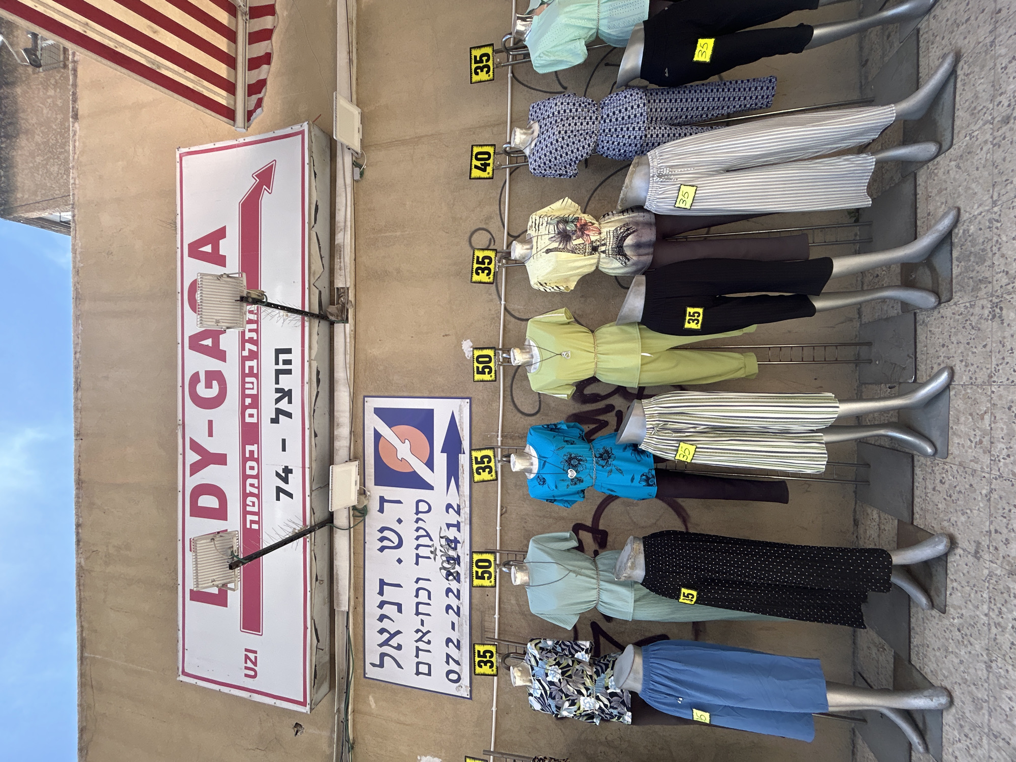 The image depicts a street-level view of a clothing store display in Rishon LeZion, Israel. A large sign with "LADY-GA" in prominent pink lettering, followed by "uzi" and Hebrew text, is mounted on a tan stucco wall. Below this sign is another rectangular sign with Hebrew text and a logo featuring a circular orange symbol with a stylized bird or airplane. Below that, a phone number "072-2224412" is visible.
In front of the wall, several mannequins are arranged, showcasing women's clothing. The mannequins are positioned along a metal rack from which prices are hung, indicated by yellow tags with black numbers. These prices include "35", "50", "40", and "35". The clothing displayed includes various tops and bottoms, such as patterned shirts, solid colored blouses and pants, polka dot skirts, and striped trousers. The store appears to be situated on a sidewalk or pedestrian area, with a cobblestone-like ground surface in the foreground. An awning with red and white stripes is visible on the upper right side of the image. The sky above is partially visible, appearing overcast. No people are actively engaged in the scene.