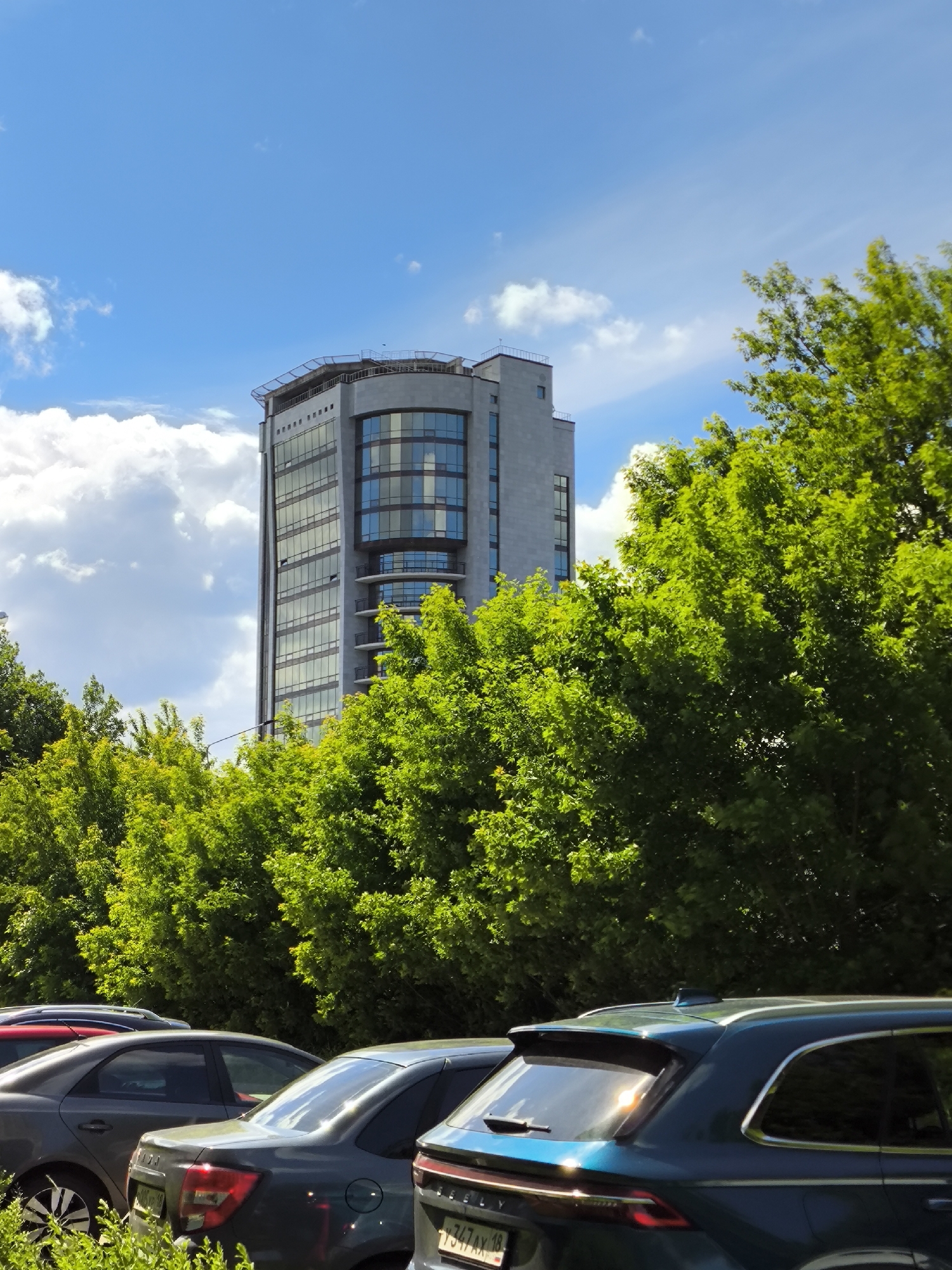 The image captures a bright, sunny daytime scene in an urban environment. A prominent, modern, multi-story building stands in the upper-middle of the frame, constructed with light grey stone or concrete panels. Its right side features a distinctive, vertically curved glass facade, while the left side has a flatter design with rectangular windows. The building's roof is equipped with a visible superstructure, including railings, and rectangular ventilation or decorative elements near the top of the flat section.
A dense band of vibrant green deciduous trees occupies the middle ground, obscuring the lower portions of the building. More trees are visible to the right of the building and in the lower foreground. The sky above is clear and blue, with scattered white cumulus clouds, particularly on the left side, and some high, wispy cirrus clouds.
In the foreground, several cars are parked, their rears facing the viewer. From left to right, a dark grey sedan is partially visible. Next to it is another dark grey sedan, identifiable by its rear lights, possibly a Lada Granta. On the far right, the rear of a dark teal or blue SUV, possibly a Geely, is visible. Its license plate clearly displays "7347AX 18", where "18" is the region code for the Udmurt Republic of Russia. No people are visible in the scene.
The location is the city of Izhevsk, Russia. The distinctive building is identified as the Business Center "Summit" (Деловой центр «Саммит»), located at ulitsa Karla Marksa, 218. The parked cars are situated in a lot adjacent to this building.