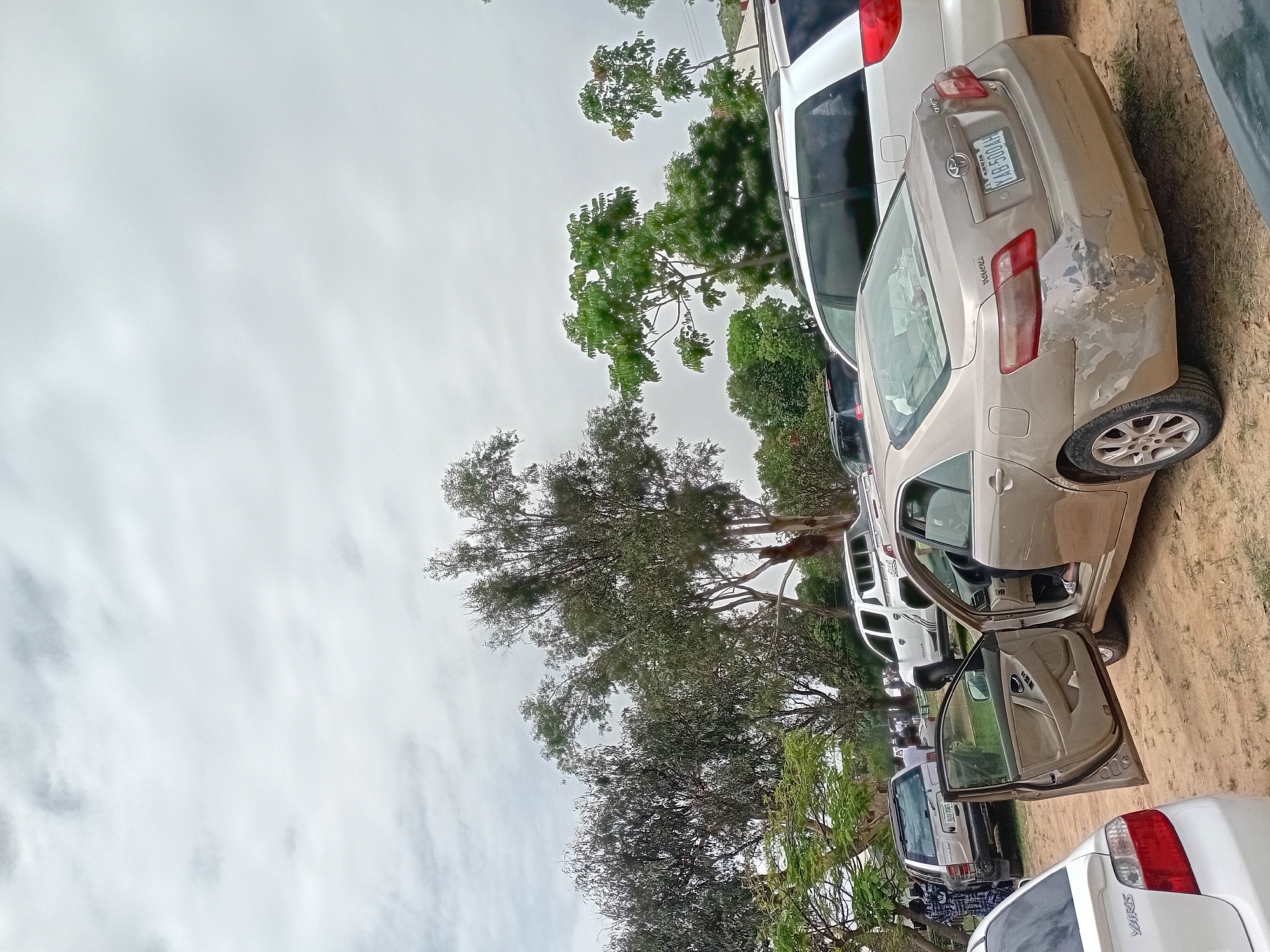 The image shows a parking area in Jos, Nigeria. Several cars are parked, including a silver Toyota Camry with an open driver's side door. The car has noticeable damage to the rear passenger side. Visible license plate number is NAS300AH. There are trees in the background, and the sky is overcast. Other parked vehicles include a white van, a white pickup, a blue car, and a white Toyota Yaris. Some people are present but blurred in the background. The ground is dirt/sand.