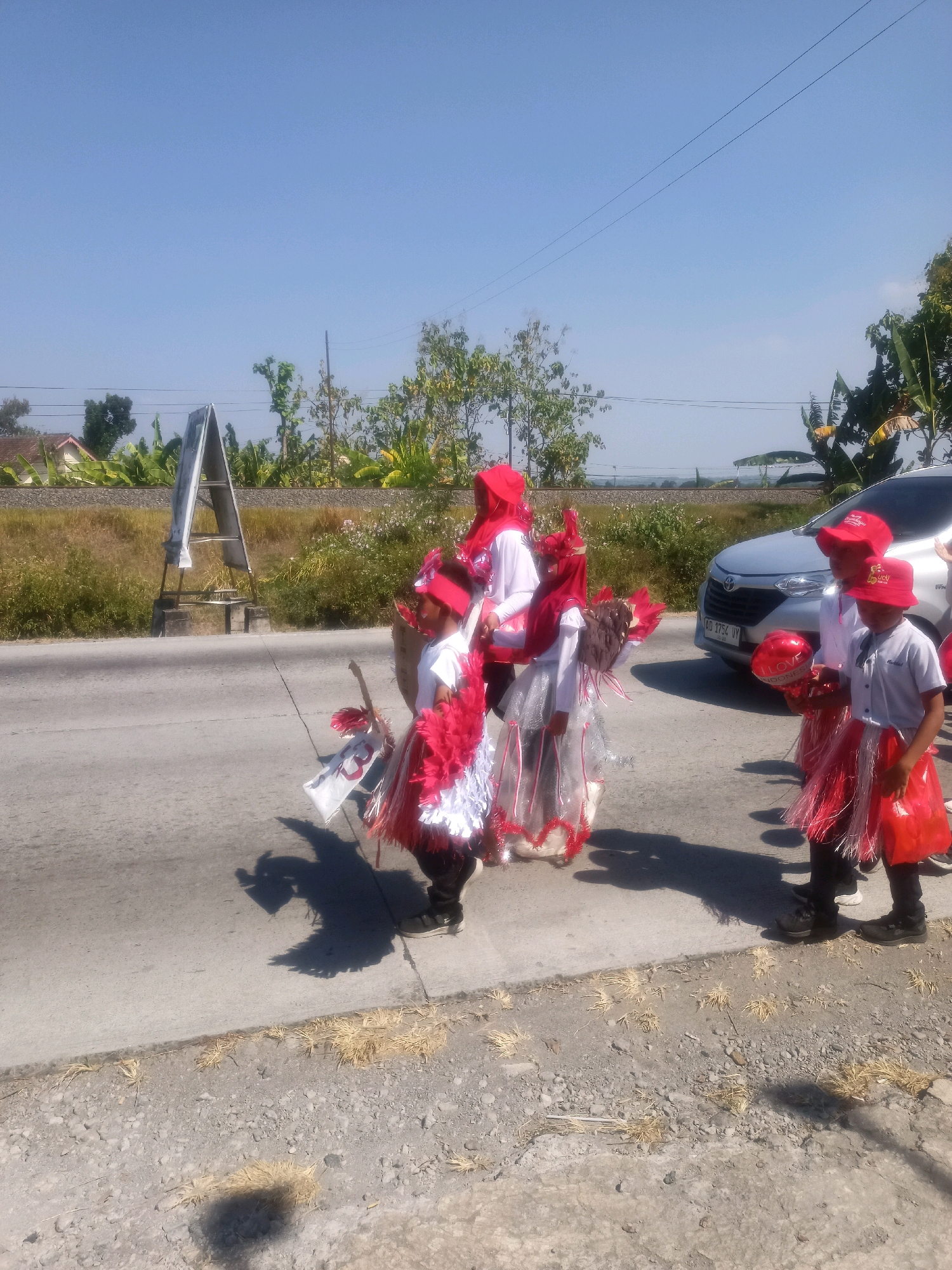 In Sumberlawang, Indonesia, a group of children in red and white costumes are participating in a procession on a sunny day. The children are dressed in elaborate outfits, possibly for a cultural event or parade, featuring red headscarves, red and white feathered decorations, and transparent skirts. One child is holding a red balloon with "I LOVE INDONESIA" written on it. They are walking along a paved road, with a silver car parked on the right side of the image. In the background, there is a railway track, lush greenery, and houses, suggesting a semi-rural or suburban setting. A triangular advertising stand is visible on the left side of the road. The shadows of the children and the advertising stand are cast on the road, indicating a bright sun.