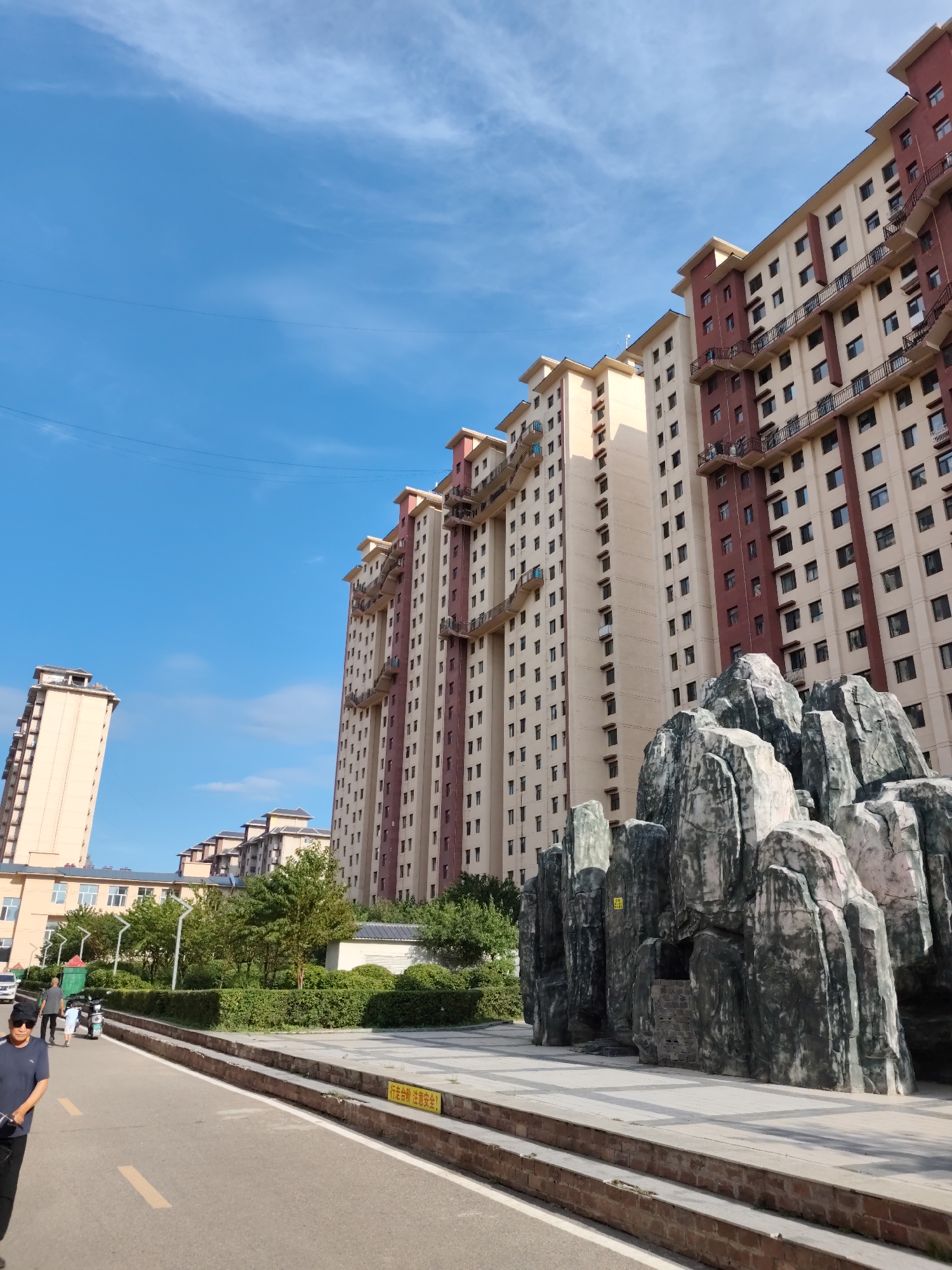 The image shows a street scene in Lindian, China, with a row of tall residential buildings on the right side, painted in alternating beige and dark red sections. The buildings have numerous windows and balconies with black railings. In the foreground to the right, there is a large, artificial rock formation with a small set of stairs leading into an opening. A paved walkway with brick edging runs in front of the rock formation and continues towards the left. On the left side of the image, there are more residential buildings and a clear blue sky with scattered white clouds. A road runs through the center foreground, with a yellow line painted down the middle. Two people are visible walking on the left side of the road, one wearing a dark shirt and sunglasses, and another person behind them pushing a scooter. A white scooter is parked on the side of the road. Trees and green bushes line the area between the road and the buildings on the left. A yellow sign with Chinese characters is visible on the walkway in front of the rock formation.