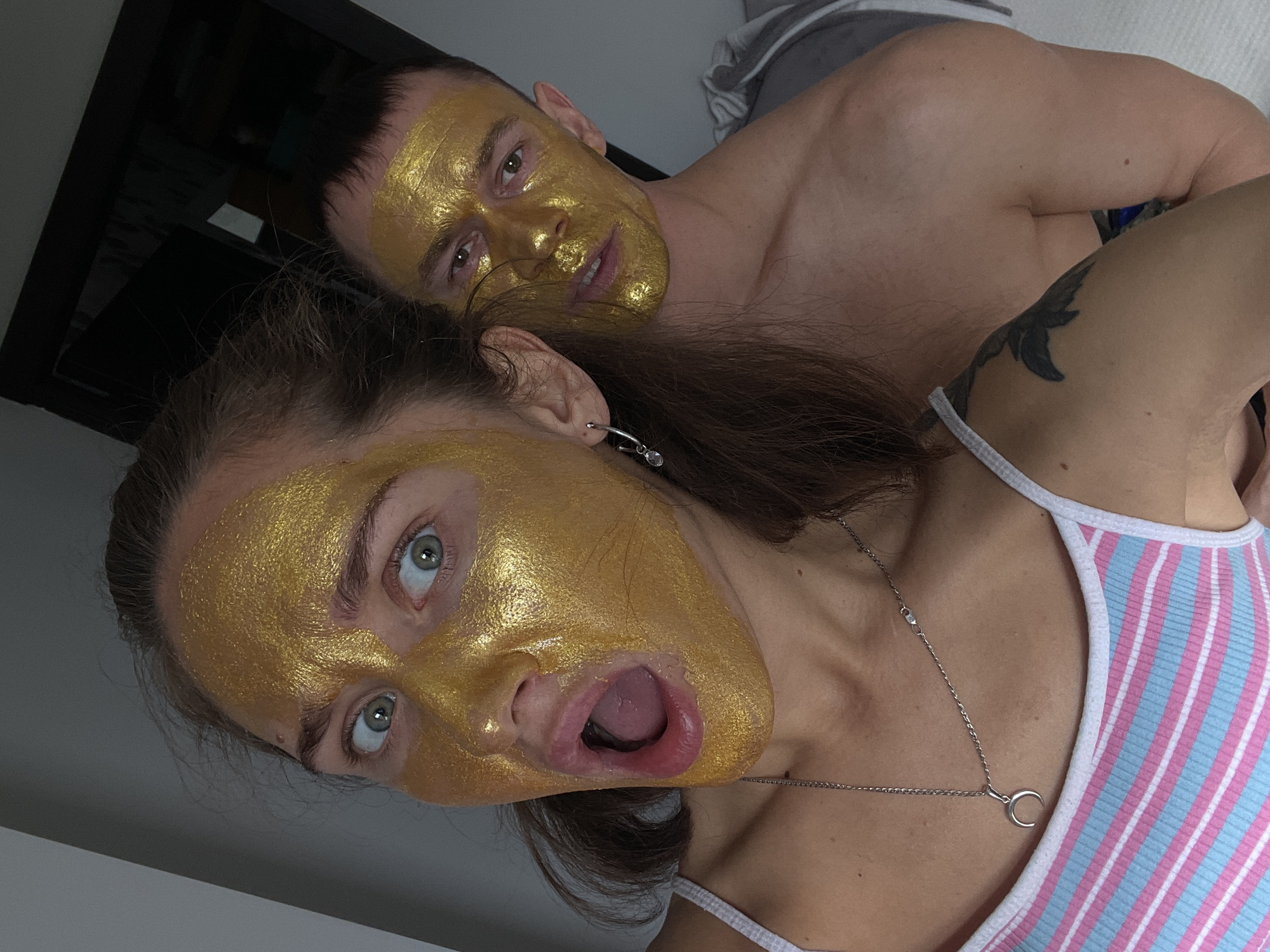Two people, a man and a woman, are posing for a selfie with golden face masks on. The woman is in the foreground, wearing a striped pink and blue top and a silver necklace with a crescent moon pendant. She has her mouth open, appearing surprised or in mock shock. The man is behind her, partially shirtless. A tattoo is visible on the woman’s shoulder. The background appears to be an indoor setting, possibly a bedroom, with a dark-colored object, which looks like a closet or part of a bed frame visible in the top left corner and a light gray surface and a striped gray and white object which might be a blanket in the top center. There is no specific visual information to confirm the image was taken in Sochi, Russia.
