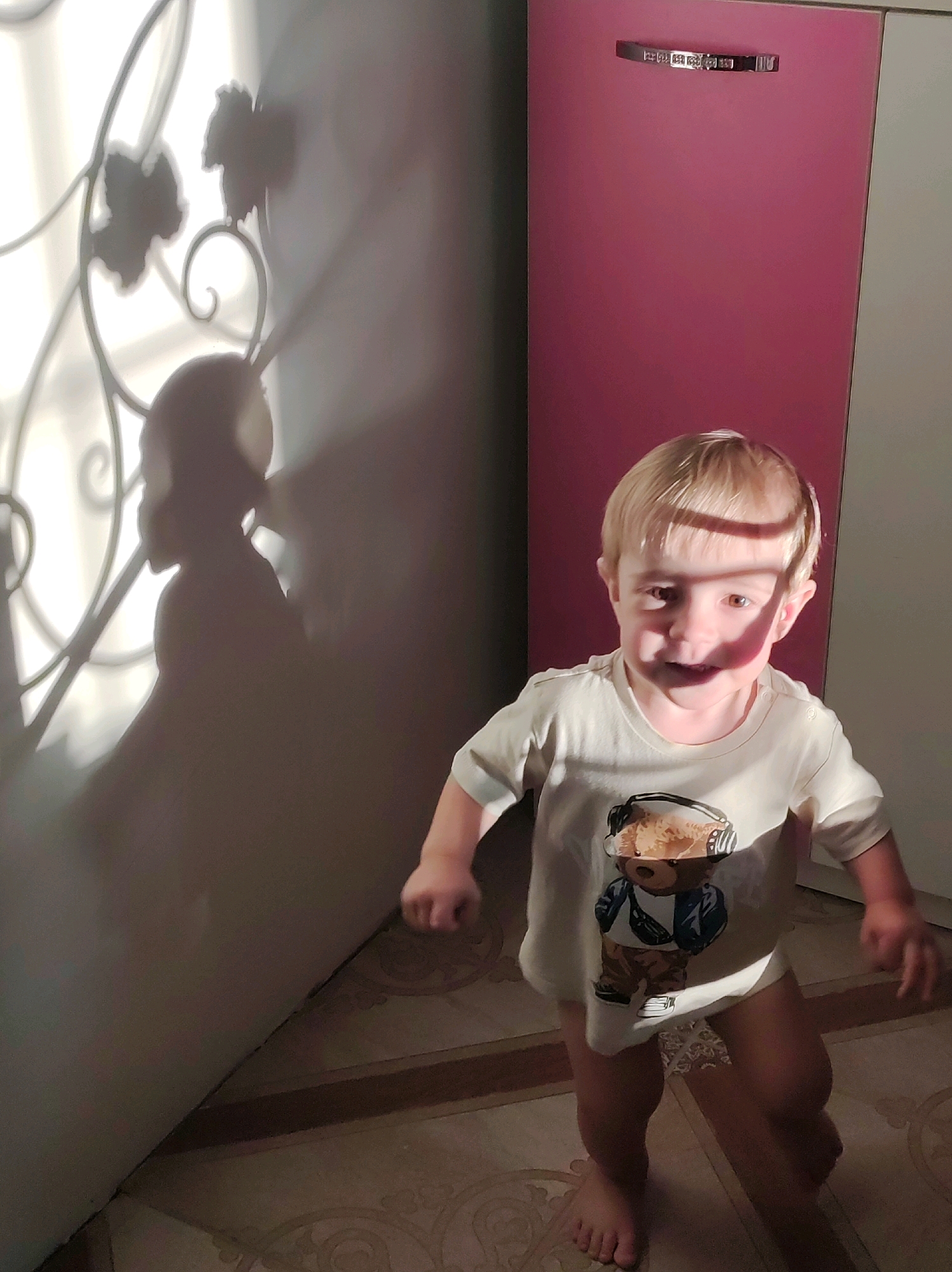 The image shows a young, blonde-haired child standing in a room. The child is wearing a light-colored t-shirt with a graphic of a bear wearing headphones and a fanny pack. The child is looking towards the camera with a slight smile. A shadow of a person is cast on the wall to the left of the child, created by a decorative metal railing. The room appears to be a residential interior, with tiled flooring and a pink cabinet or closet visible in the background. There is bright light entering from the left, creating strong shadows. The location is identified as Fokinsky District, Bryansk Oblast, Russia.