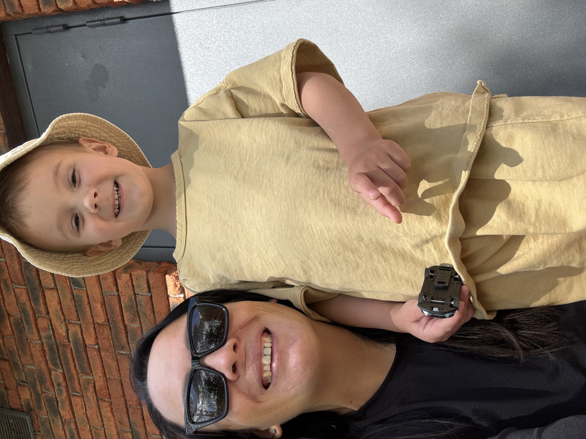 A woman and a child are in the foreground, smiling at the camera. The woman is wearing sunglasses and a black shirt, while the child is wearing a straw hat and a yellow t-shirt. The child is holding a small toy car. The background shows a brick wall and a gray door. The image appears to be taken outdoors on a sunny day. The orientation of the image is rotated.