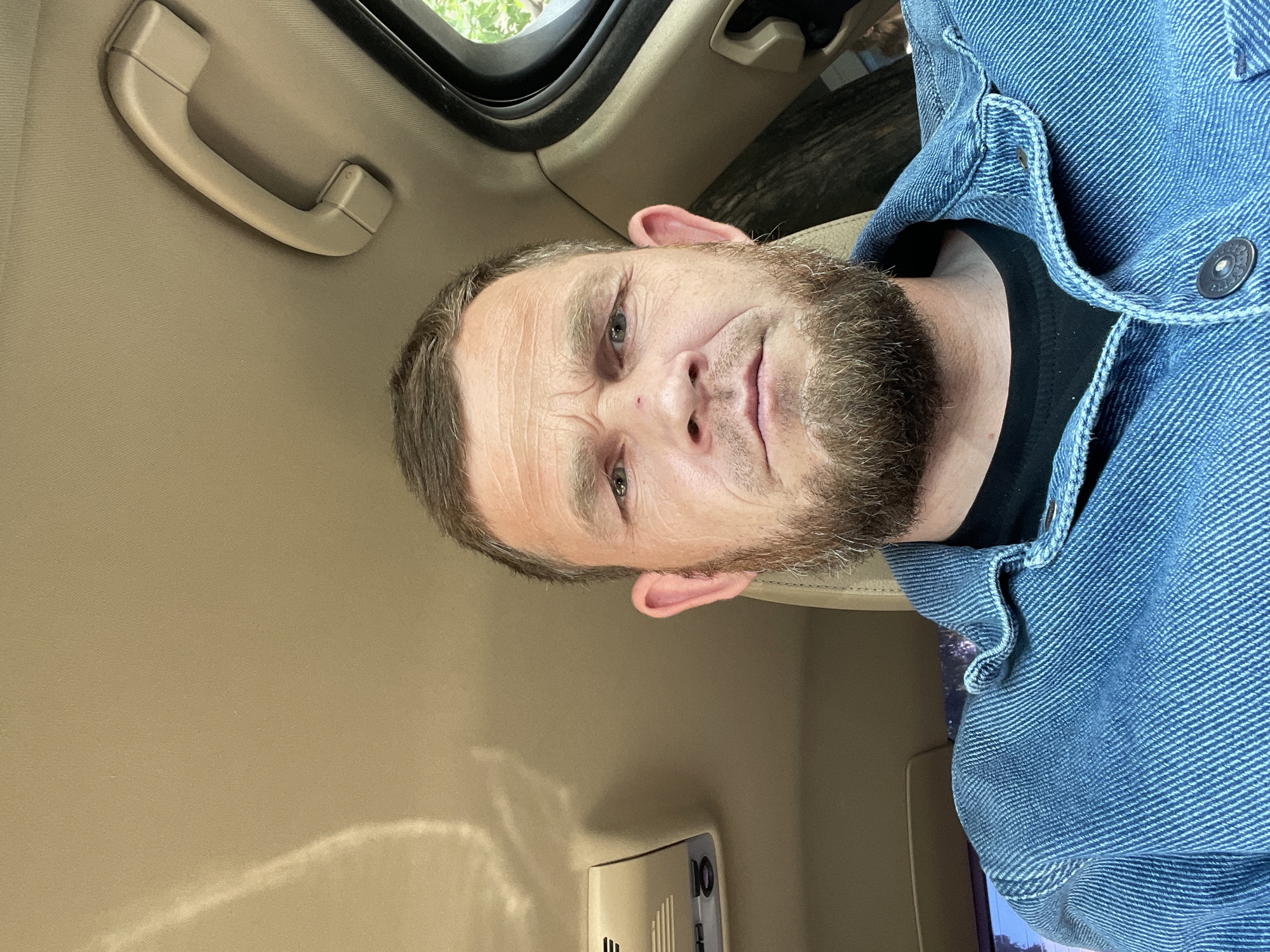 A man with short brown hair, a beard, and light-colored eyes is looking at the camera. He is wearing a black t-shirt and a blue denim jacket. He appears to be inside a vehicle, judging by the interior elements visible, such as a beige headliner, a handle, a sunroof, and a dashboard element. The location within the city of Krasnodar, Russia is not evident from the image alone.