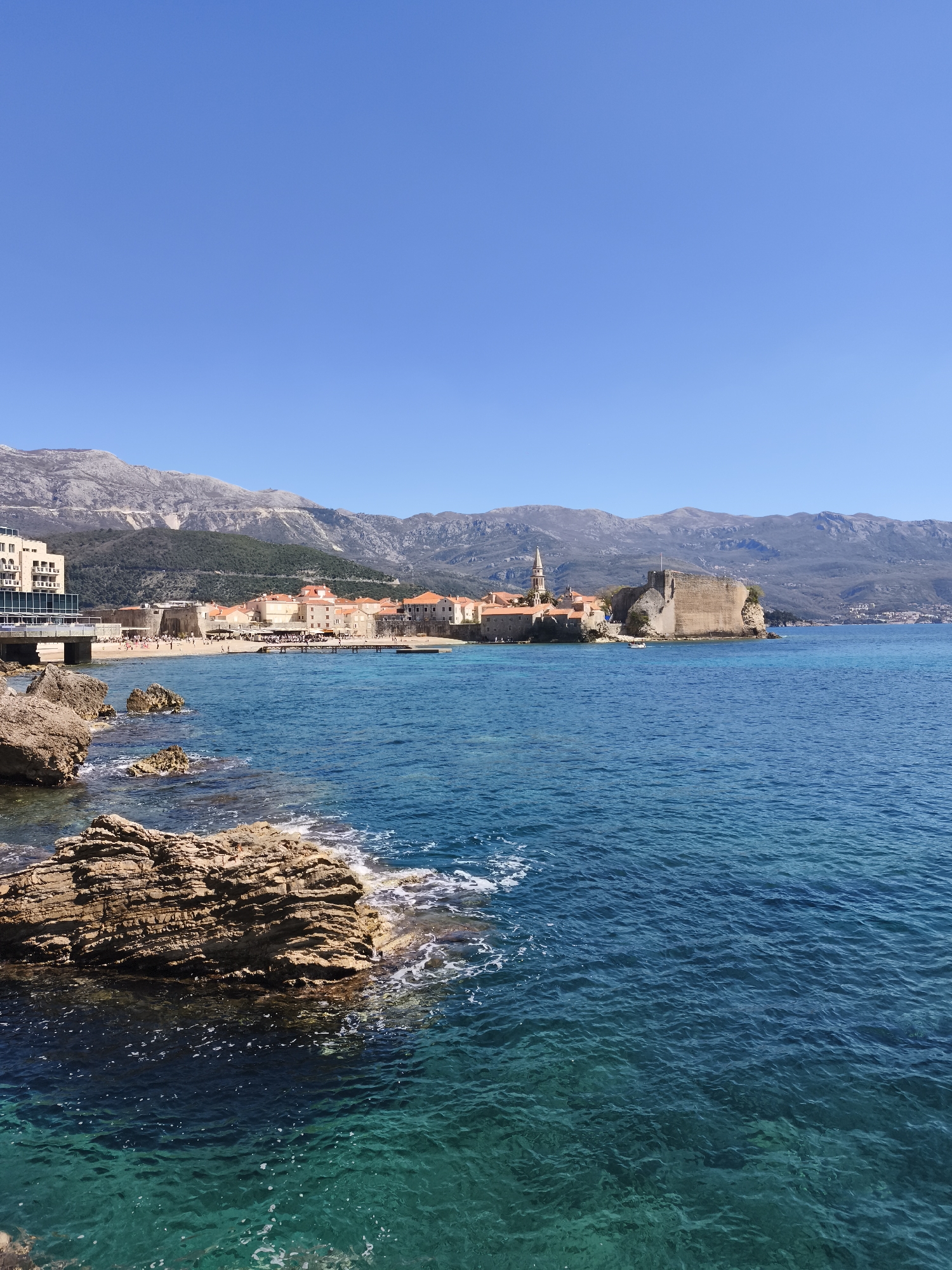 The image depicts a panoramic view of Budva, Montenegro, captured on a clear day under a bright blue sky. The foreground is dominated by turquoise-colored water. Rocky outcroppings jut out from the sea, their layered surfaces exposed to the sun. Beyond the water, a coastline is visible with a sandy beach where people are gathered. A building complex stands out along the shore. Behind the buildings is a range of mountains with various peaks. In the center of the view is Budva Old Town, with its distinctive red-tiled roofs and a tall church steeple. A stone fortress is located at the edge of the old town.