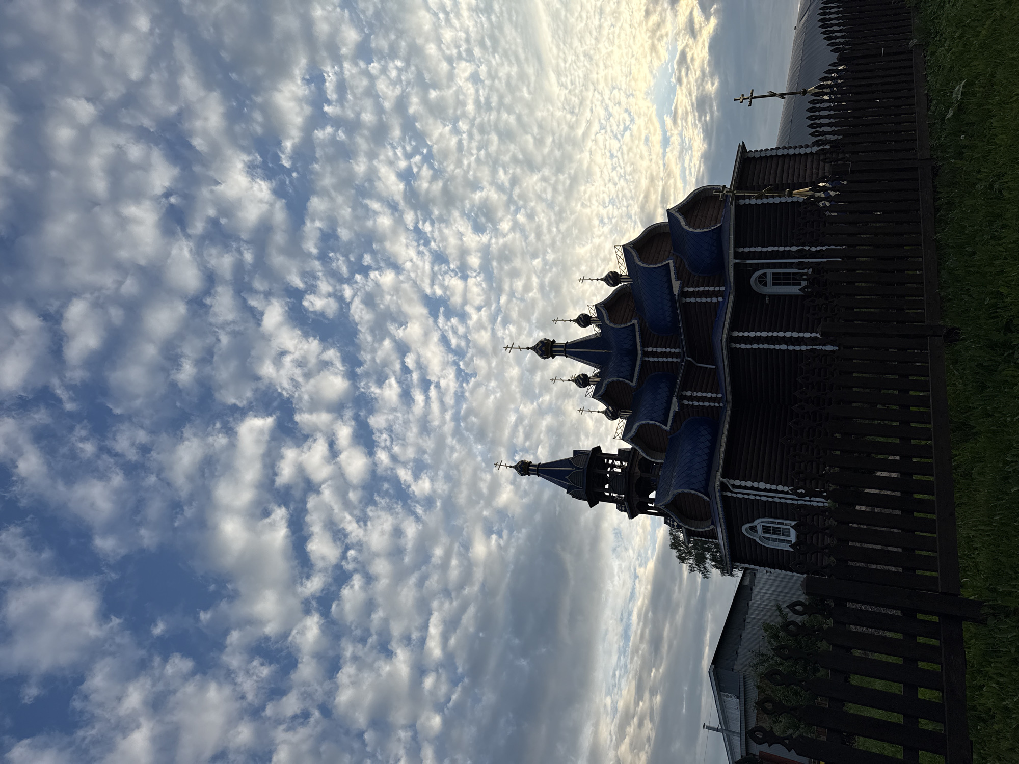 The image shows a wooden Orthodox church with blue roofs and multiple onion domes, located in the village of Kukui, Russia. A dark wooden fence runs across the foreground, with green grass and some plants visible behind it. To the left of the church, a less ornate building with a metal roof is partially visible. The sky above is filled with white, textured clouds against a blue background. The overall scene depicts a rural religious structure under a dramatic sky.