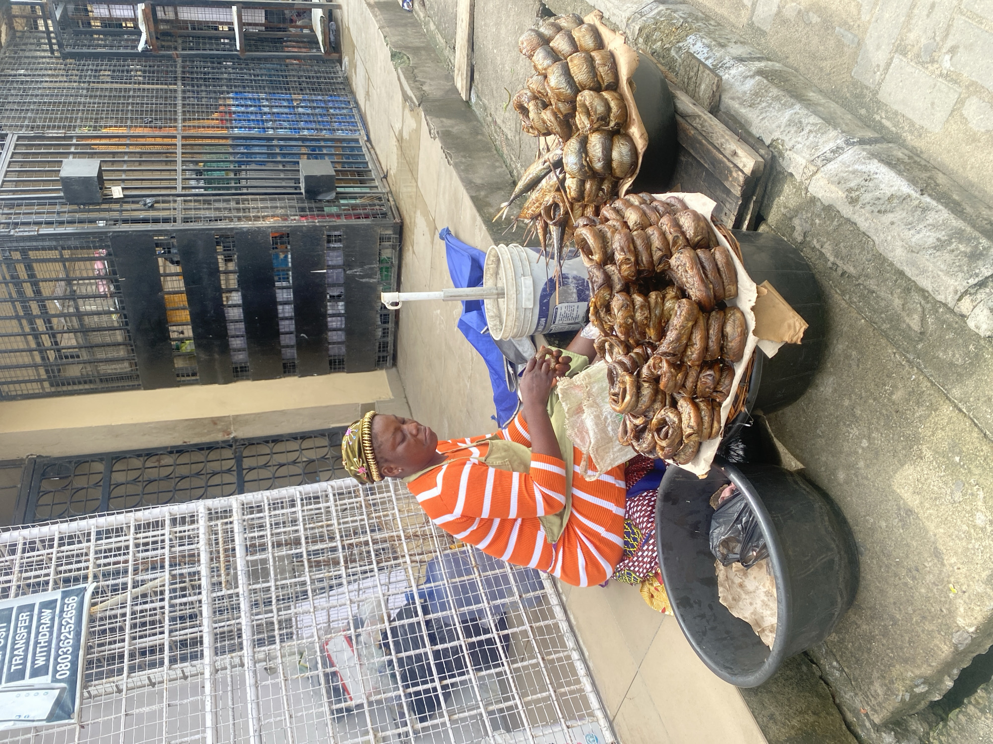 An overhead medium shot captures an outdoor market stall in an urban setting. A woman, wearing an orange and white striped dress and a patterned headwrap, sits on the ground. She is facing to her right, and her hands are engaged in sorting through items. In front of her, arranged in two large black bowls, are piles of smoked fish. The stall is set up on a concrete pavement, with a large metal grate structure and a building visible in the background. The overall scene depicts a vendor preparing her goods for sale.