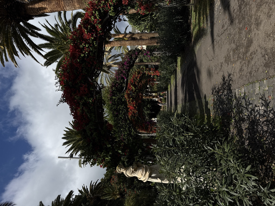 The scene depicts a paved pathway within Parque García Sanabria, Santa Cruz De Tenerife, Spain, under a sky with scattered white and grey clouds and visible patches of blue. In the left foreground, a light-colored stone statue of a draped female figure on a pedestal is partially obscured by dense green foliage. The dark paved pathway, bordered by a strip of cobblestones on its left, recedes into the distance, leading beneath a series of interconnected metal arches heavily covered with vibrant red and some purple bougainvillea flowers. Further down the path, in the mid-ground, several indistinct human figures are visible, appearing to walk or stand. The environment is lush with various green shrubs and numerous tall palm trees, some prominently positioned on the right and in the background. Sunlight casts sharp, defined shadows from the surrounding vegetation onto the pathway and ground surfaces.