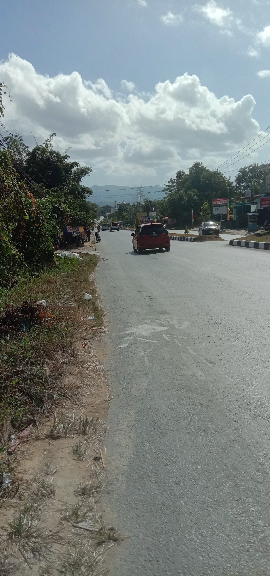 The image shows a street scene in Kendari, Indonesia. The foreground features a paved road with a rough asphalt surface. The shoulder of the road on the left side is unpaved, with dry grass and some scattered debris, including plastic bags and other refuse. A dense cluster of trees and bushes lines the left side of the road, with some exposed branches and vines.
On the road, a red car is visible in the middle distance, traveling away from the viewer. Its license plate reads "SE 971 GA". Further down the road, several other vehicles are driving in the same direction. The road appears to be a multi-lane highway.
To the right of the road, there are several commercial establishments. One building has a sign that reads "TOKO" (shop in Indonesian) and another sign with "BSI" and "ATM" indicating a bank. There are also power lines and poles along the side of the road. In the background, hills and mountains are visible under a partly cloudy sky with large, white cumulus clouds. A few people can be seen on the left side of the road near the trees, possibly near a roadside stall or gathering. A motorcycle is parked on the side of the road.