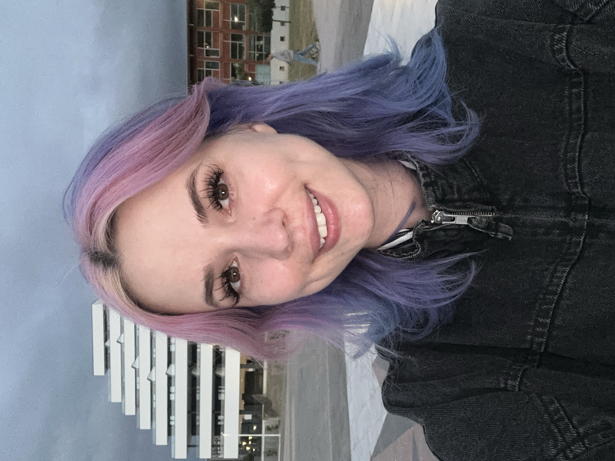 This is a selfie of a young woman smiling broadly, her head tilted slightly. She has distinctive two-toned hair, transitioning from a light pinkish-purple on her left side to a deeper blueish-purple on her right, styled in loose waves. Her brown eyes are framed by long, dark eyelashes and well-defined eyebrows. She is wearing a dark, likely black or dark grey, denim jacket with a prominent silver zipper visible on the front.
The background indicates an urban environment, consistent with Gijón, Spain. To the left, a modern building with a facade composed of white vertical slats is partially visible. Further back and to the right, a multi-story building constructed of reddish-brown brick with numerous windows can be seen. The sky is overcast or cloudy, suggesting either late afternoon or early evening light conditions. The woman's smile conveys a cheerful and contented emotion. No visible text is present in the image.