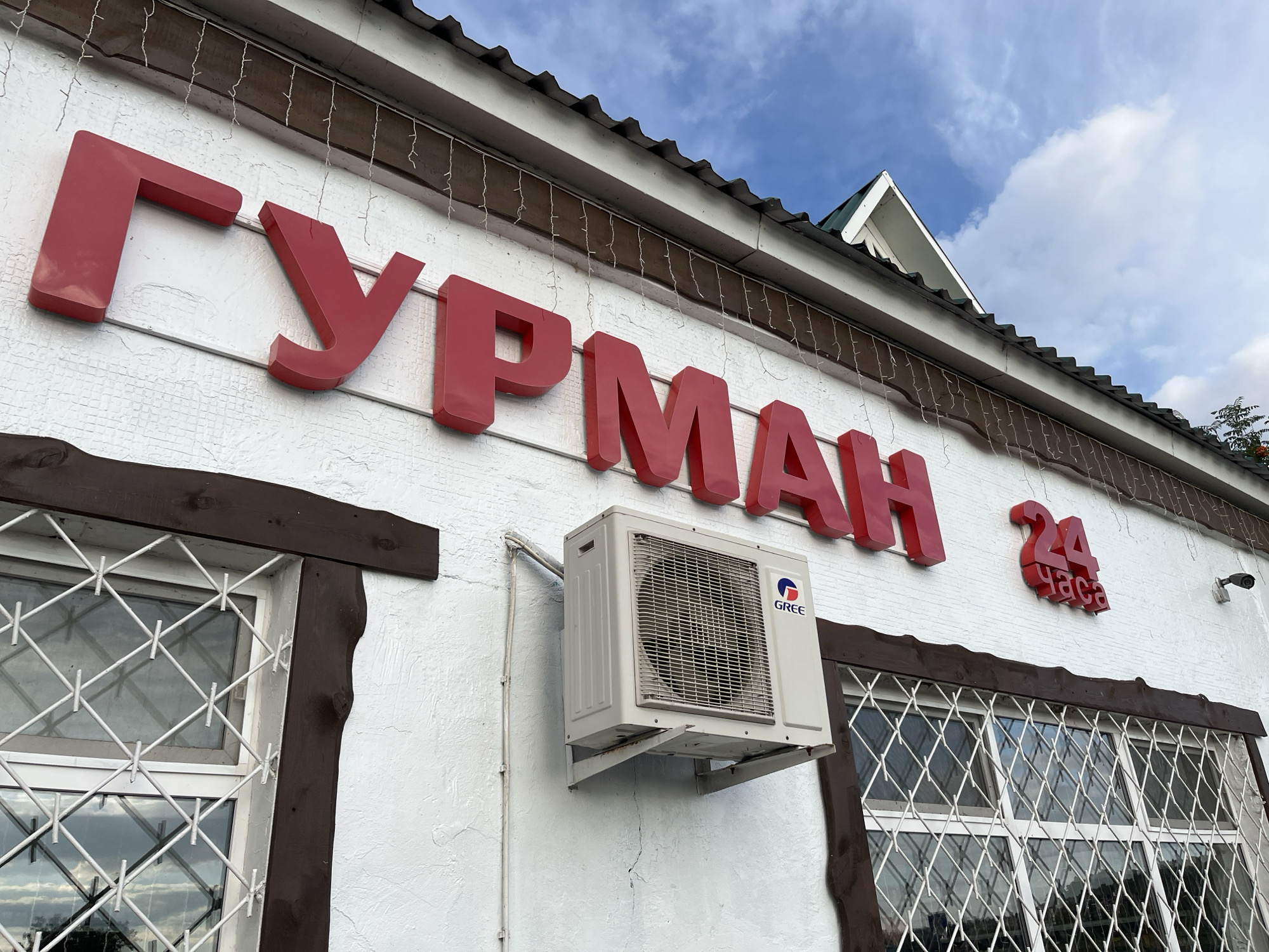 The image shows a close-up of a white building's facade with large red lettering spelling out "ГУРМАН 24 ЧАСА". This translates to "GOURMET 24 HOURS", indicating a 24-hour establishment. The building has a dark brown trim around two windows, one of which is visible on the left and partially on the right. Both windows have white decorative metal grates. An outdoor air conditioning unit, branded "GREE", is mounted on the wall between the lettering and the right window. A security camera is visible mounted on the wall above the right window. String lights are draped across the building above the lettering. The sky above is blue with some white clouds. The building appears to be located in the city of Blagoveshchensk, Russia.