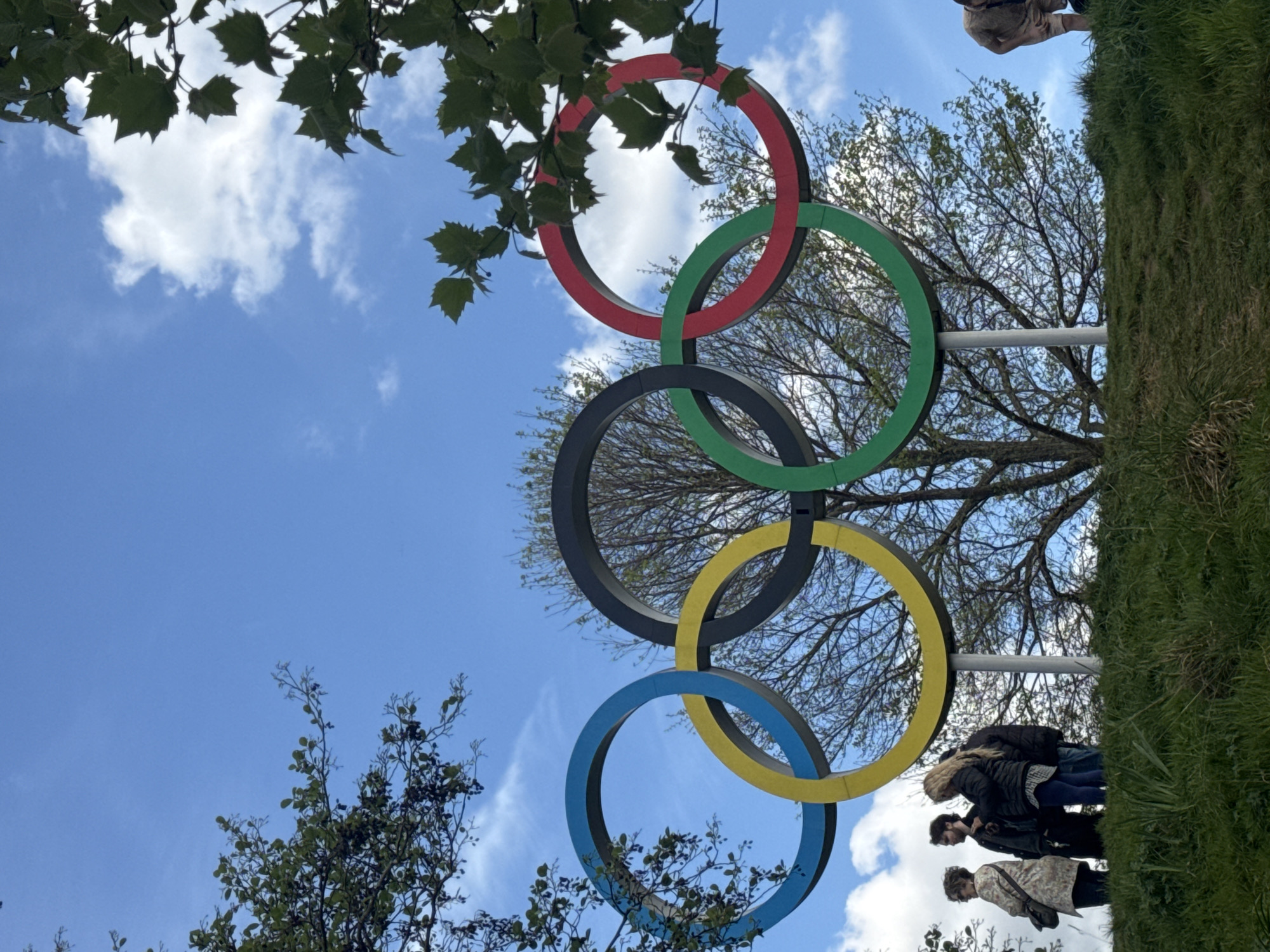 The image shows the Olympic rings monument in London, United Kingdom, angled upwards against a bright blue sky dotted with scattered clouds. The rings, colored red, green, black, yellow, and blue, are arranged in their traditional overlapping pattern. The rings are framed by trees, some with lush green leaves and others with bare branches. In the background, a grassy hill is visible, with a few people standing on it near the monument. The perspective is from below, looking up at the rings, emphasizing their size and presence against the sky.