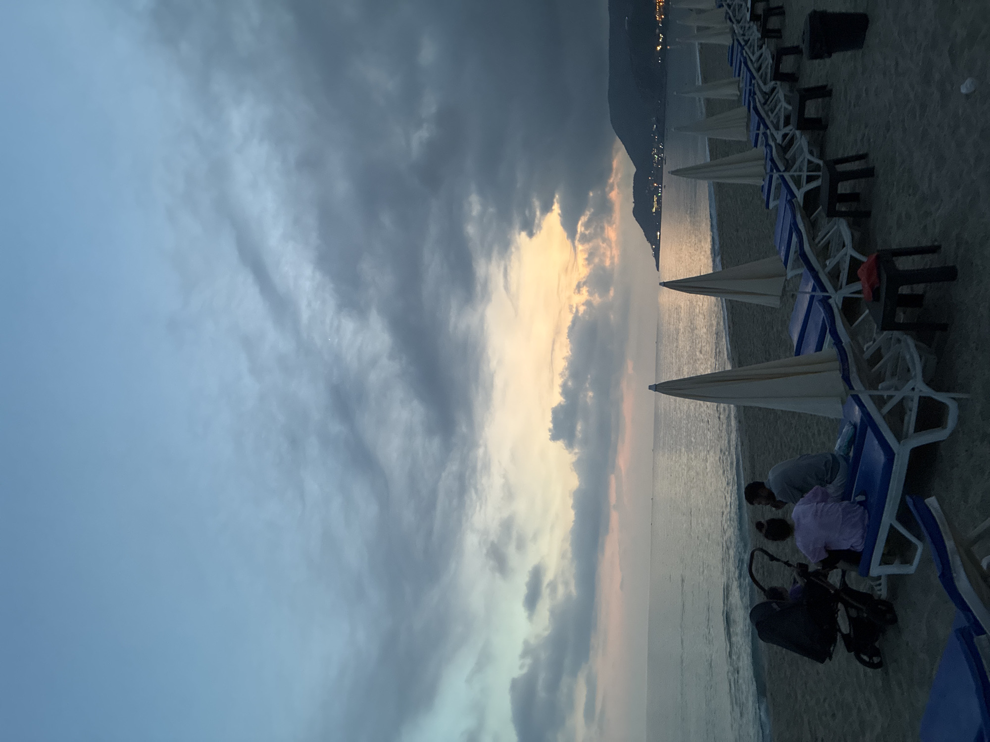 A family of three, including two adults and one child, are seated on blue loungers on a sandy beach in Alanya, Turkey. The adults are facing away from the camera, with one adult holding the child. A stroller is parked next to the loungers. Several closed beach umbrellas are arranged in rows behind them. The sea is visible in the background, with a gently sloping shoreline. The sky is overcast with dramatic clouds, tinged with hues of orange and pink from the setting sun. The silhouette of a coastal town with lights is visible on a hill in the distance. The overall impression is one of a tranquil evening on the beach.