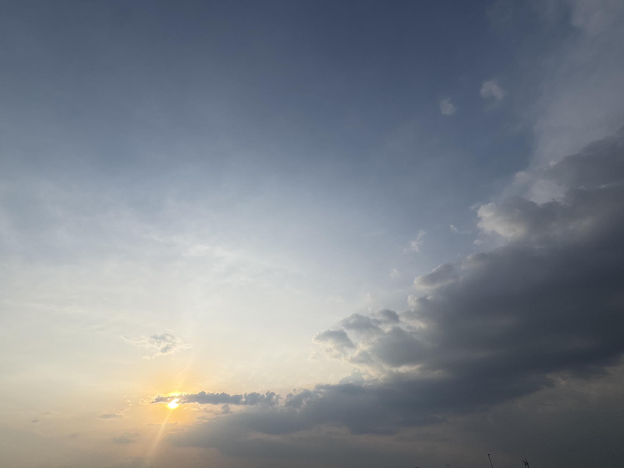The image shows a daytime sky in San Francisco Ocotlán (Ocotlán), Mexico. The sky transitions from a light blue in the upper left corner to a darker, cloudier area in the lower right corner. The sun is visible in the lower left, partially obscured by clouds, with sun rays emanating from it. The sky contains a mix of thin, wispy clouds and larger, more dense clouds. The horizon is barely visible at the very bottom of the image.