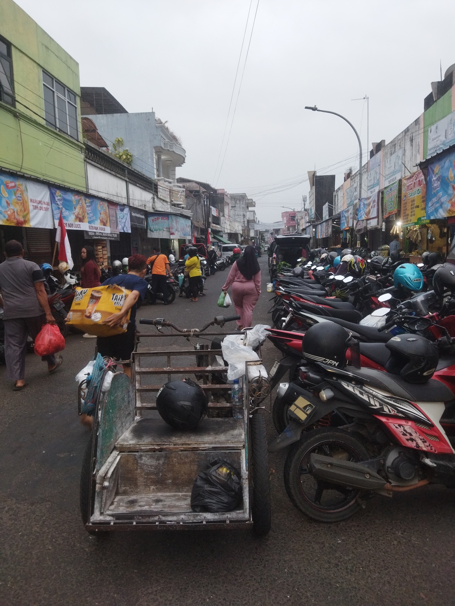 The image depicts a street scene in Banjar, Indonesia, likely a market area, characterized by a narrow street lined with businesses and parked motorcycles. In the foreground, a two-wheeled cart with a wooden cargo area is positioned on the left side of the street. A black helmet and a black plastic bag are visible in the cart.
Several people are present in the scene. On the left, a man wearing a gray shirt and pants is walking away from the camera, carrying a red plastic bag. A woman in a maroon top and dark pants is standing near the cart. In the middle ground, a person in a navy blue shirt and dark shorts is carrying a large yellow box. To the right, a woman in a light pink outfit is walking away from the camera, carrying a white plastic bag. Other individuals are seen in the background, some near parked motorcycles.
The street is densely populated with parked motorcycles of various colors, predominantly red and black. Many of these motorcycles have helmets placed on them. The buildings on either side of the street appear to be shops or market stalls, adorned with numerous banners and advertisements, some featuring text in Indonesian. The sky is overcast, suggesting a cloudy day. The overall atmosphere is that of a busy, possibly commercial, urban environment in Banjar.