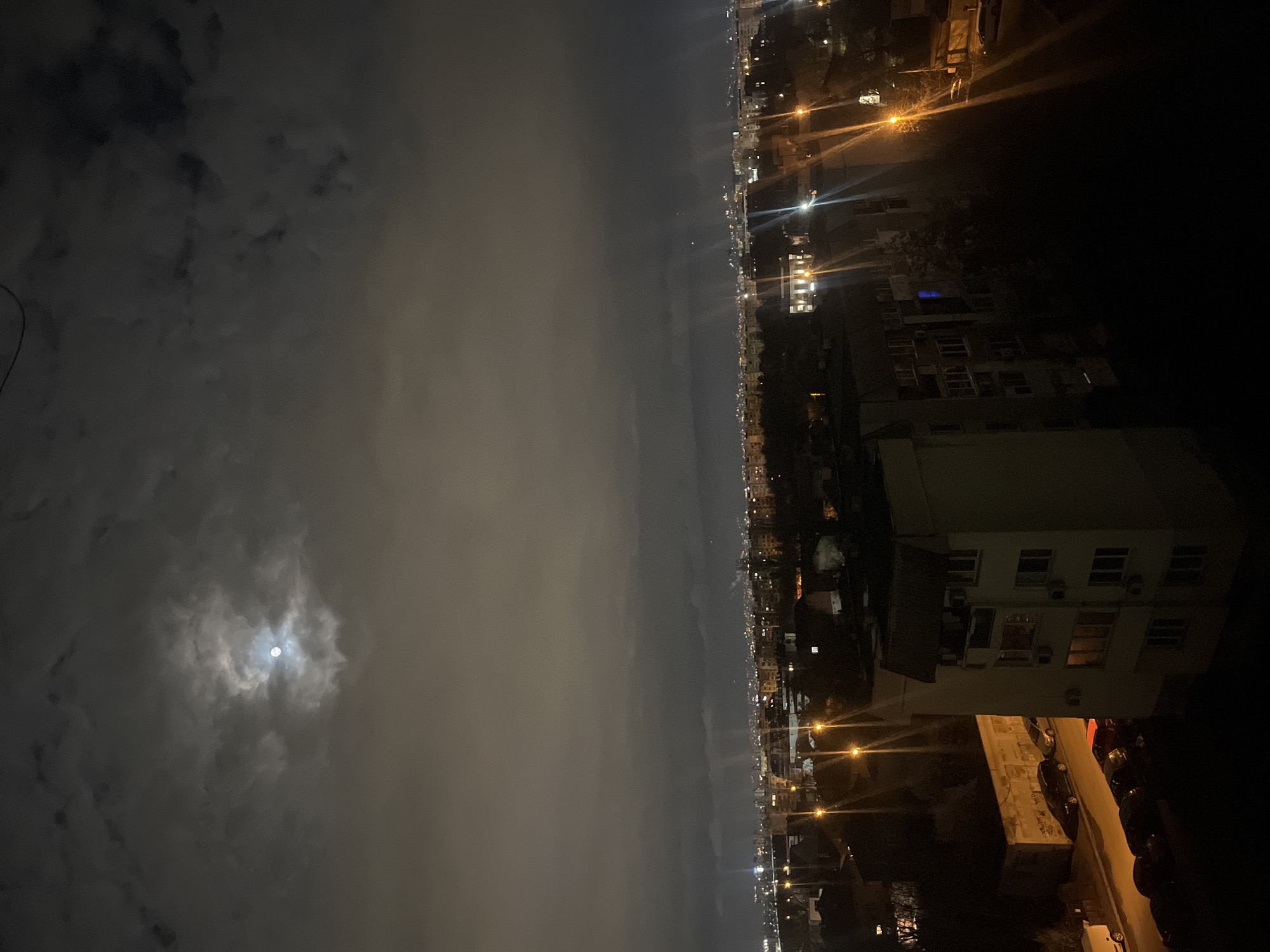 The image shows a night view over a city, likely Sofia, Bulgaria. The sky is cloudy, with the moon visible through a break in the clouds on the left side. On the right, there are buildings with illuminated windows and streetlights. Cars are parked along a road next to one of the buildings. The image appears to be taken from an elevated position, possibly a high floor of another building.