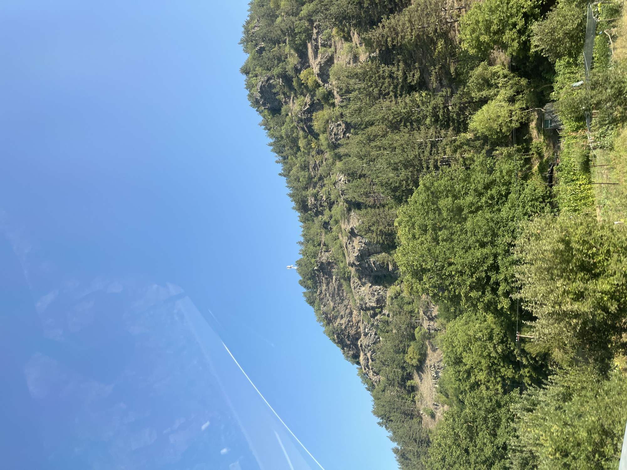 The image captures a hillside covered in lush green trees and rocky outcrops, under a clear blue sky. A white tower or antenna structure is visible near the summit of the hill, perched on one of the rocky sections. The foreground shows dense foliage and vegetation, suggesting a rural or natural setting. There are no people visible in the image. The scene appears to be a natural landscape with a man-made structure on the hillside. This description corresponds to a view of the landscape in the vicinity of Krklja, North Macedonia.