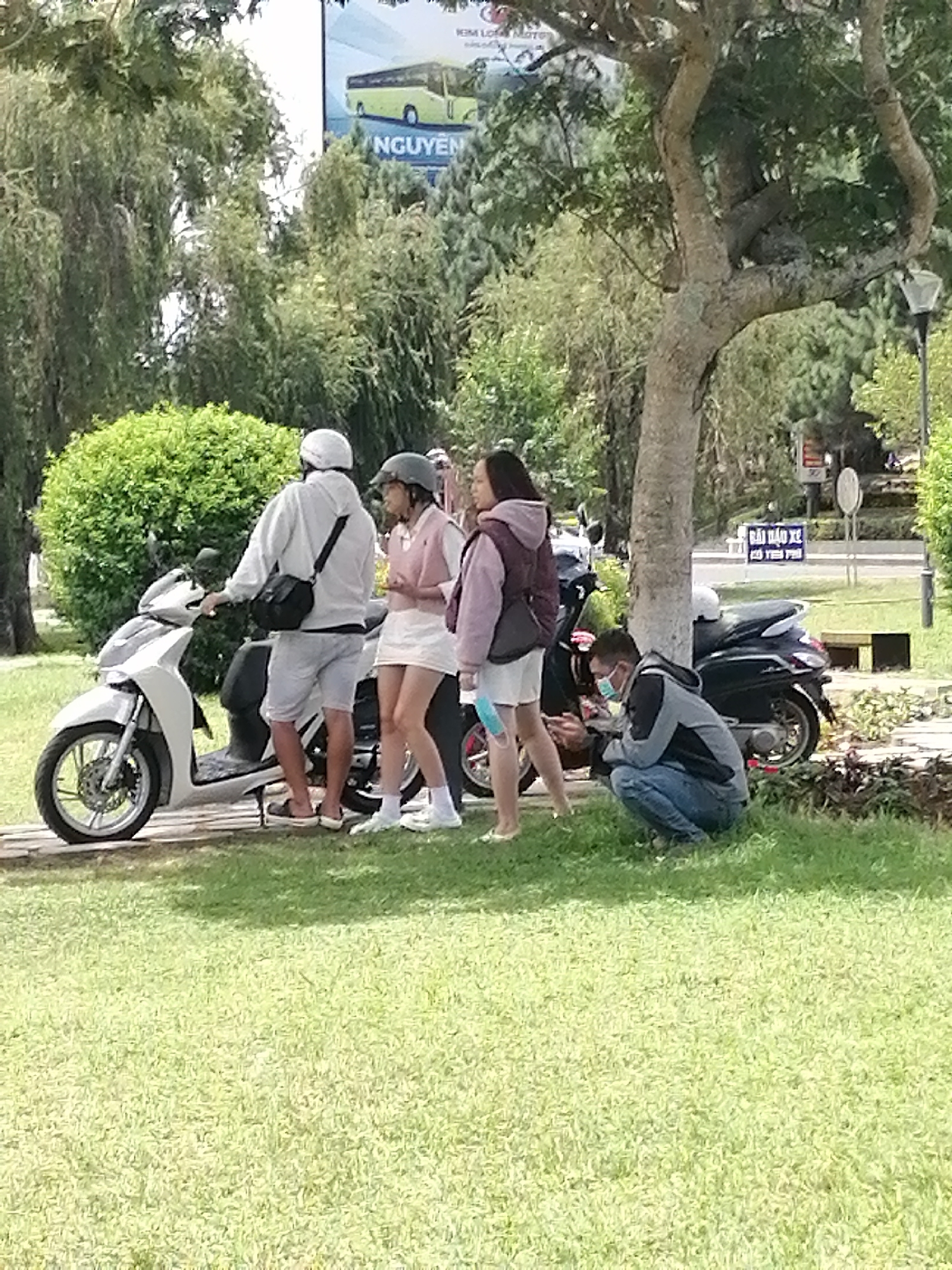 In Da Lat, Vietnam, on a sunny day, two young women and a young man are gathered near a white scooter. The woman on the left, wearing a white hoodie, gray shorts, and flip-flops, stands by the scooter. Next to her, a woman in a pink polo shirt and white skirt, and another woman in a light purple vest and white skirt, are engaged in conversation. A man in a blue and gray jacket, blue jeans, and a face mask is crouched on the grass, looking at his phone. A black scooter is parked behind them. The scene is set in a park with lush green grass, trees, and a bright blue sky. A billboard in the background advertises a bus tour.