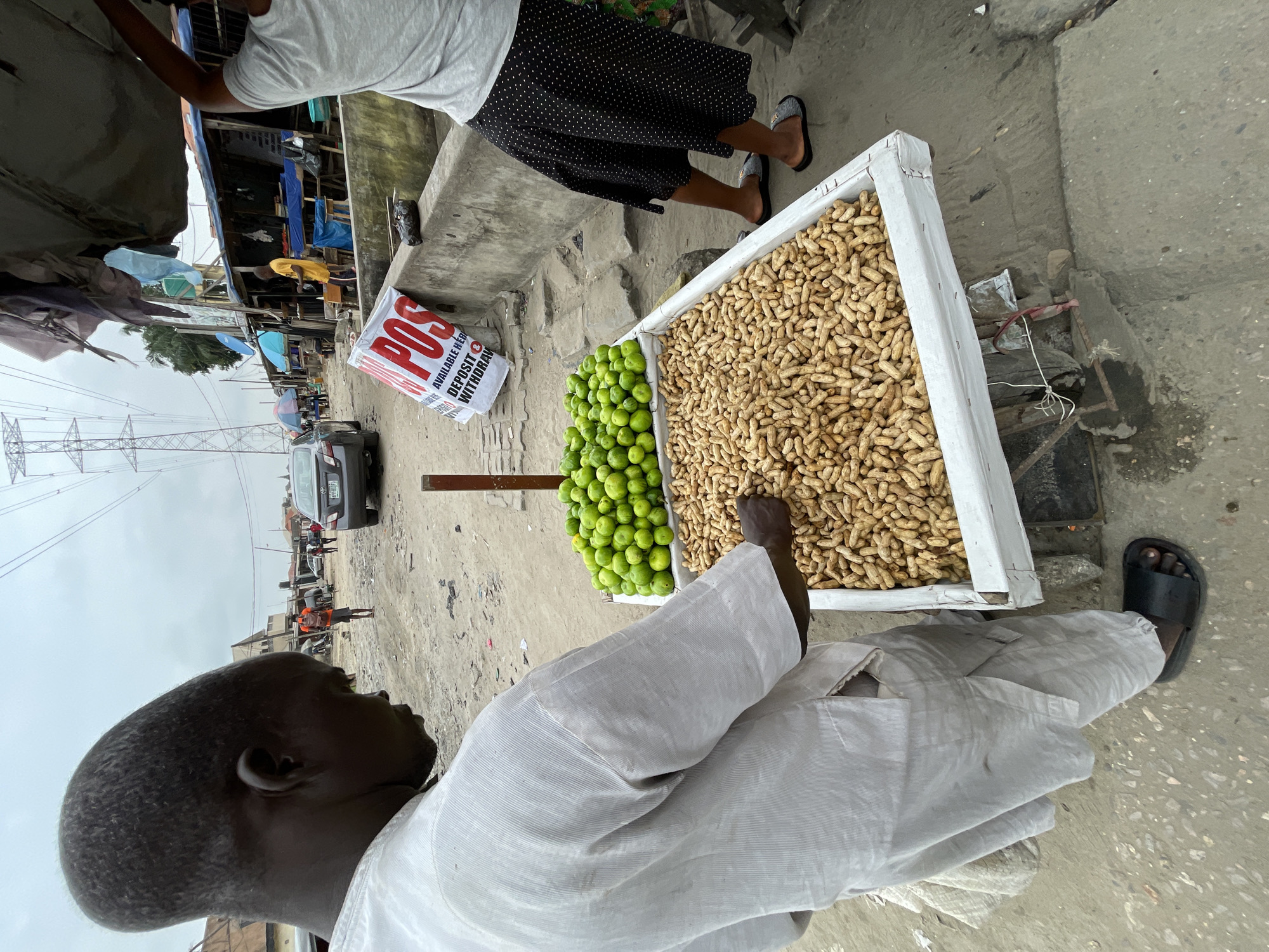 A man with a pronounced, elongated cranium stands to the left of a vendor's cart in Ado, Nigeria. He wears a loose-fitting, light-colored tunic and trousers. His right hand is extended towards the cart, fingers touching a pile of peanuts. The cart, a white wooden box on a metal frame, is laden with peanuts in shells and a smaller section filled with green limes. In the background, a busy street scene unfolds with stalls, a car, and other pedestrians. A large electricity pylon is visible in the distance, and a white banner with red lettering, possibly advertising a financial service, hangs from a nearby structure. The ground is dusty and uneven.