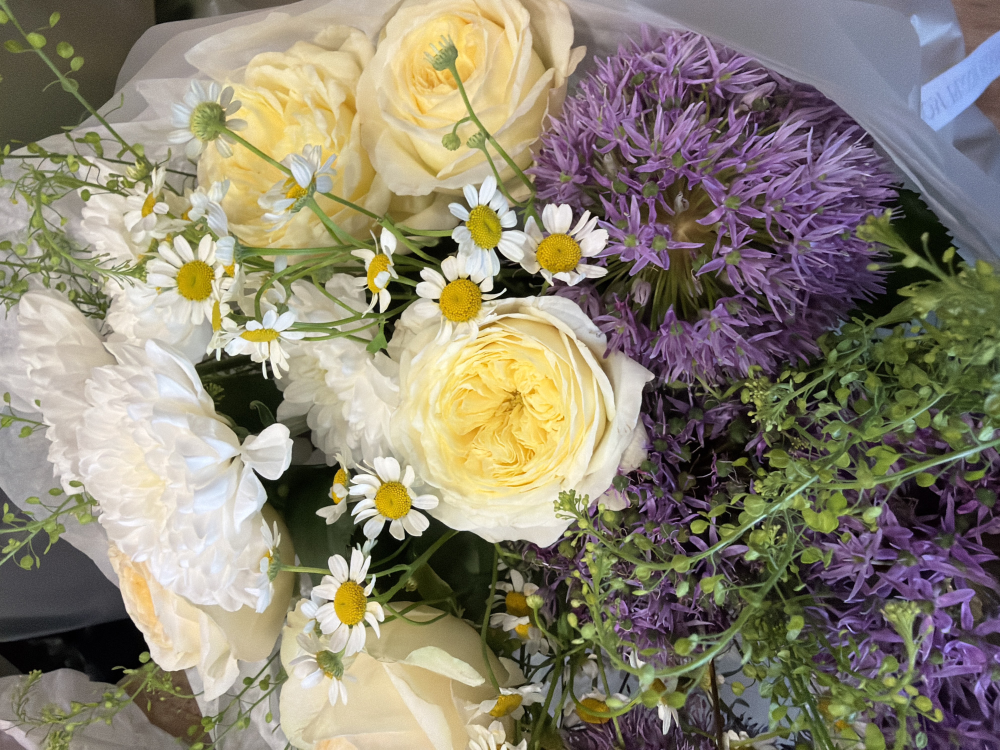The image is a close-up, overhead shot of a bouquet of flowers. The bouquet contains a variety of flowers, including large white chrysanthemum-like blooms, creamy yellow roses, small white daisies with yellow centers, and spiky purple allium flowers. There are also delicate green sprigs of foliage interspersed throughout. The flowers are wrapped in white paper or cellophane. The lighting is bright and natural, highlighting the textures and colors of the blooms. There are no people or discernible urban environmental elements in the image.
