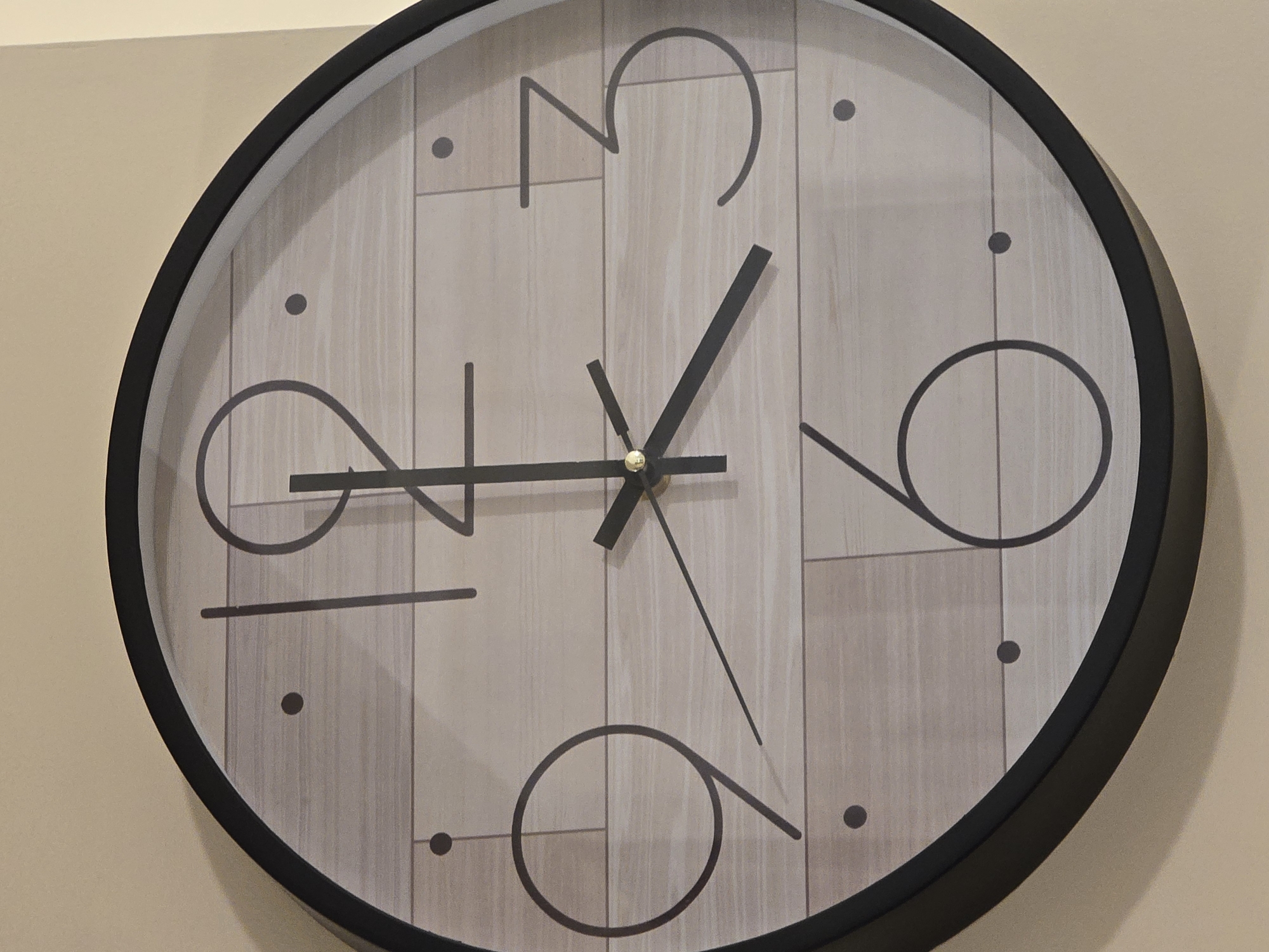 Here is a close-up shot of a round wall clock. The clock has a black outer frame and a light-colored face with a wood grain pattern. The numbers 3, 6, 9, and 12 are represented with stylized, minimalist line drawings. The minute markers are indicated by small dots. The hands of the clock show the time as approximately 1:31. The clock is hanging on a light beige or off-white wall.