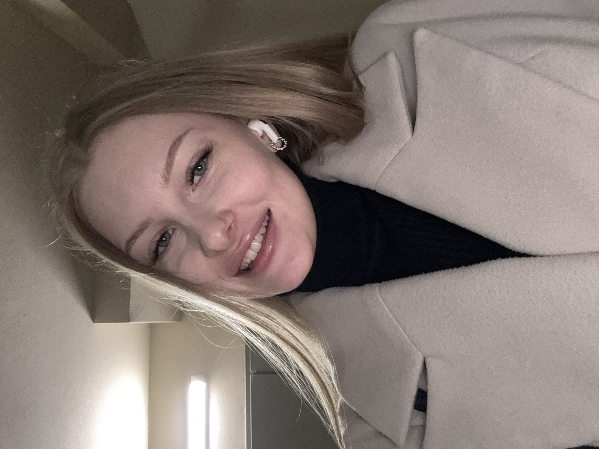 A young woman is shown in a selfie, slightly angled, wearing a black turtleneck sweater and a light beige coat. She has blonde hair and is wearing a wireless earbud in her left ear. The background shows a pale wall and a rectangular light fixture. It is not possible to ascertain whether the location is in Perm, Russia from this image.