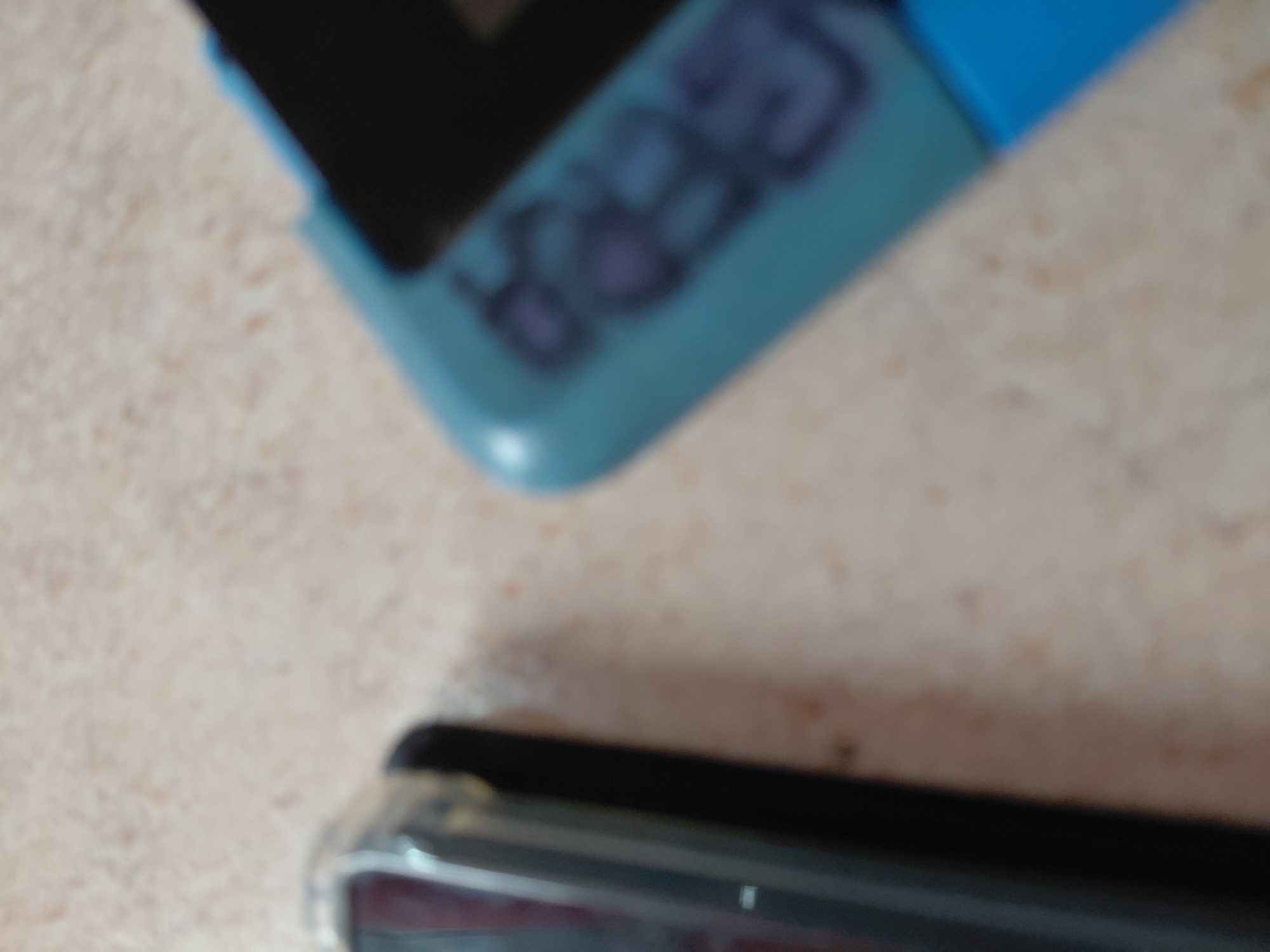 The image captures a blurry, close-up view of two electronic devices resting on a light-colored, textured surface, likely indoors. No people are visible, and no specific activity is taking place; the items appear stationary.
In the upper left portion of the frame, a turquoise or teal-colored handheld device is prominently featured. It has a dark, rectangular area that could be a screen or a dark casing, and to its right, several distinct, dark purple or blue button-like shapes are visible. Due to the significant blur, any markings or text on these buttons are unreadable. The top right edge of this device has a brighter blue segment.
Towards the bottom right, partially entering the frame, is a black electronic device, possibly a smartphone, encased within a clear or translucent protective cover. Only a small, indistinct portion of this object is visible.
The background surface appears beige or light tan with a faint, mottled texture. The overall image is severely out of focus, rendering specific details, including any potential text or brand names, completely illegible. The time of day and weather cannot be determined from the provided visual information, nor are there any specific cues to suggest the location of Santa Ana, Venezuela.